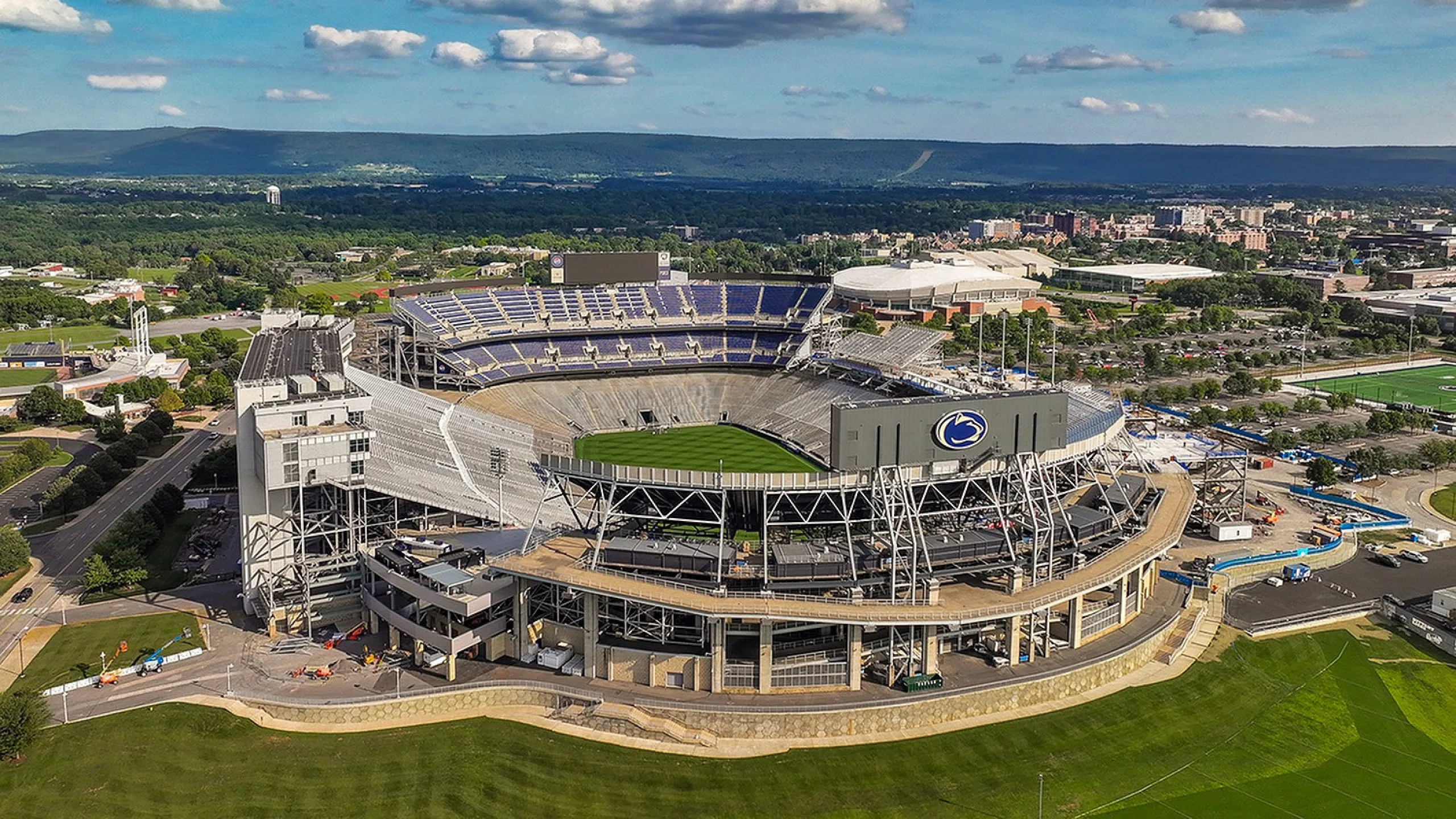 Aerial view of a vibrant stadium amidst lush green surroundings, featuring a large seating capacity and striking architectural design under a clear blue sky.
