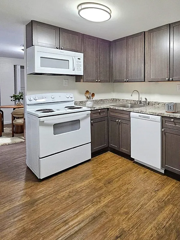 Modern kitchen with sleek dark wood cabinets, granite countertops, and essential appliances adjacent to cozy dining area with warm wooden flooring.
