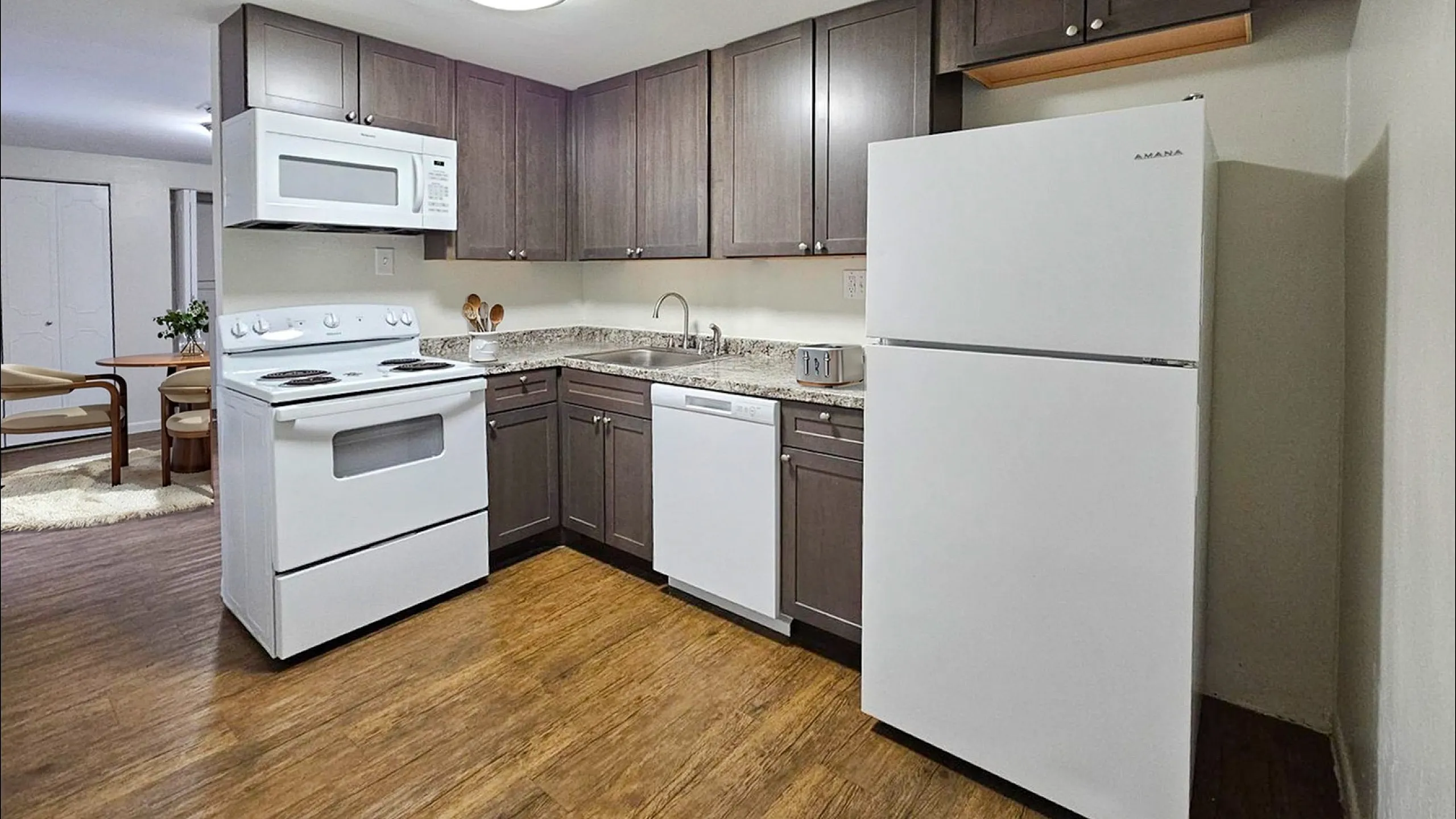 Modern kitchen with sleek dark cabinets, white appliances, and wooden flooring, opens to a cozy dining area.