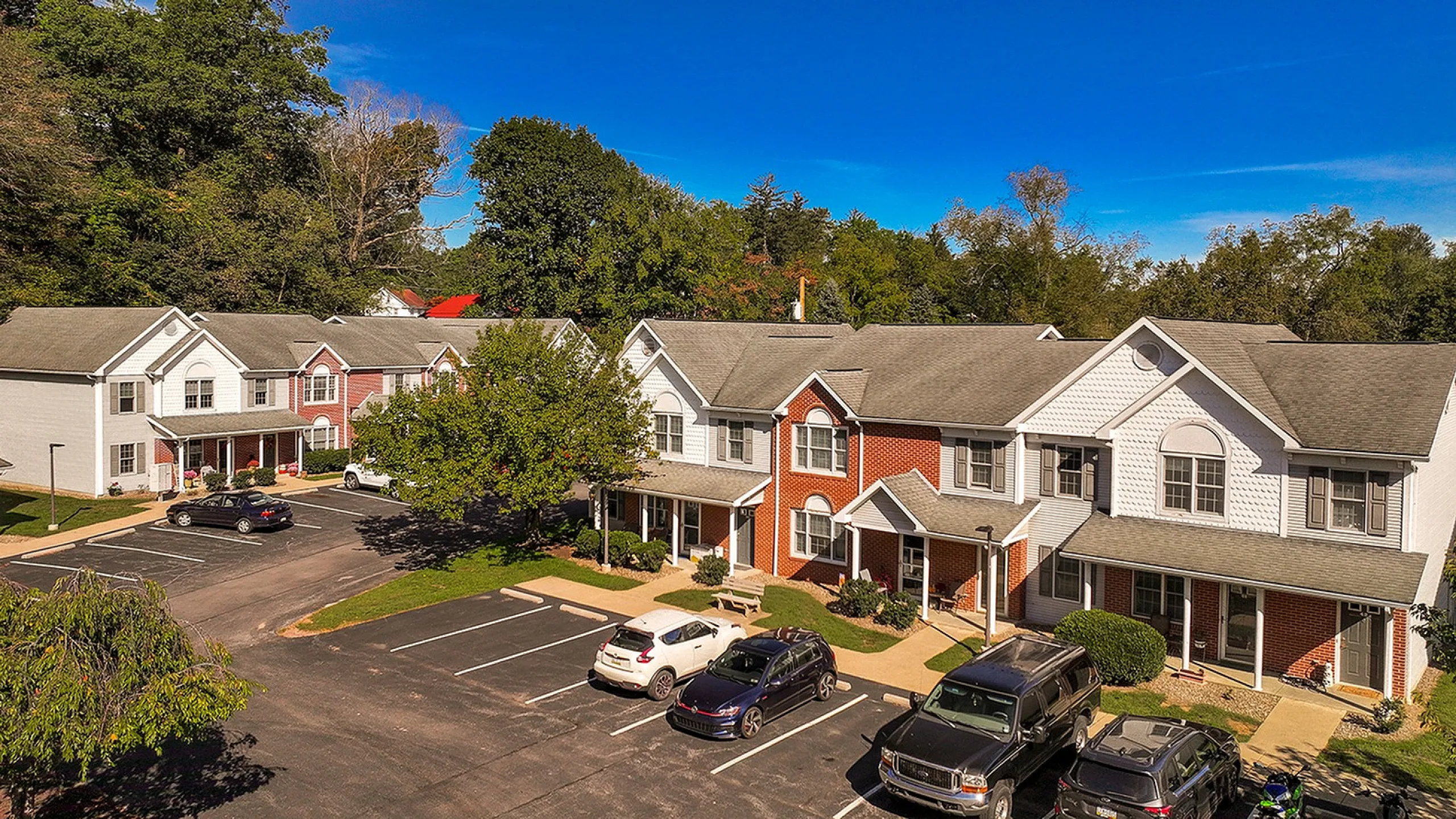 Charming townhouse complex surrounded by lush greenery, featuring ample parking and inviting front porches under a clear blue sky.
