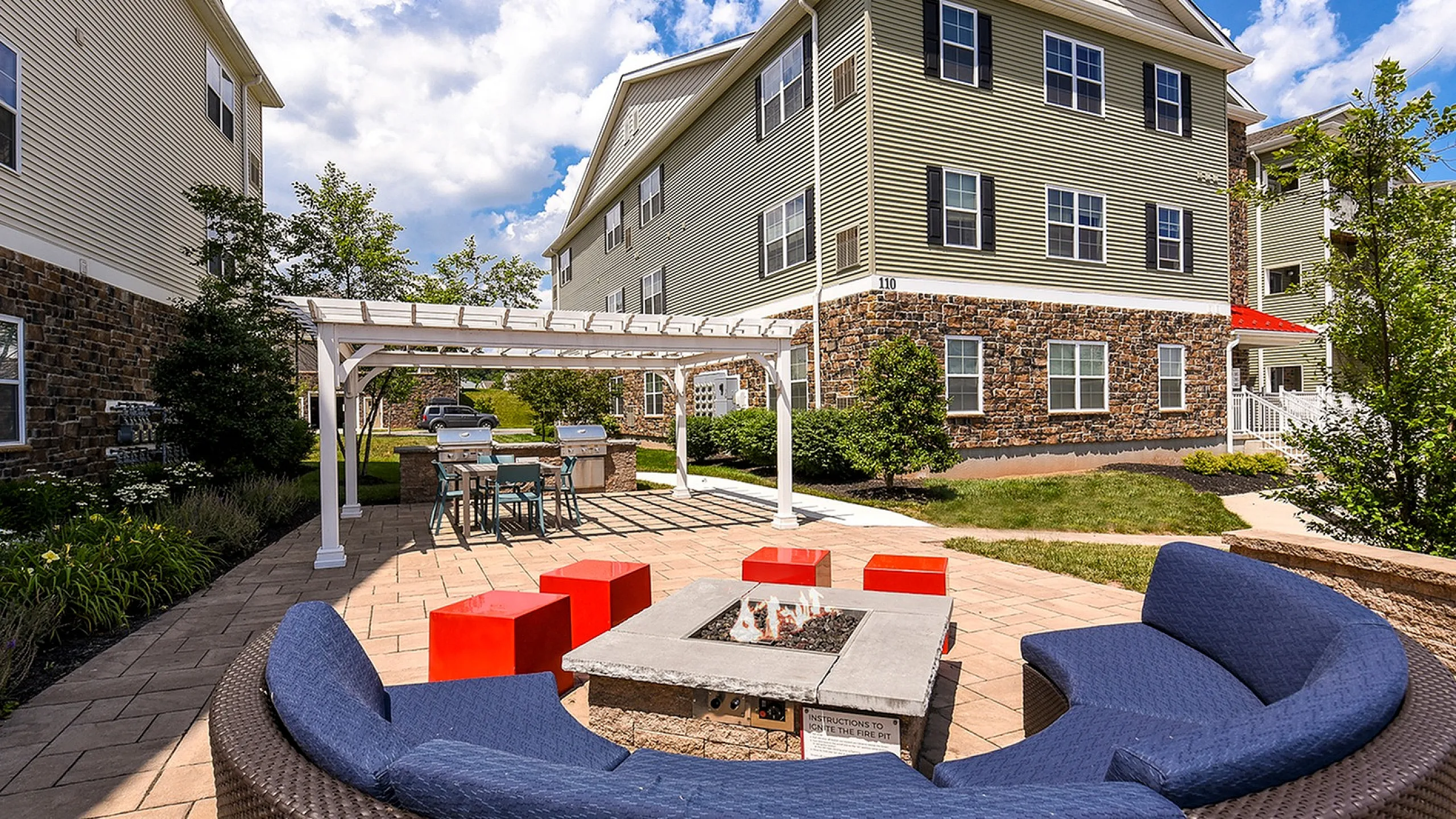 Charming outdoor space featuring a cozy fire pit with stylish seating, adjacent to a modern apartment building with a welcoming pergola and grill area.