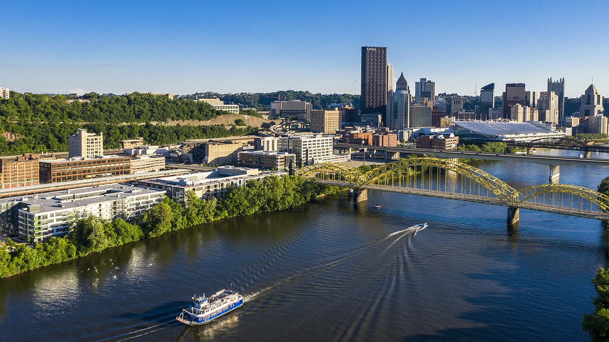 Breathtaking aerial view showcasing a vibrant cityscape with modern architecture, green spaces, and a bustling river complete with a scenic bridge and boats, perfect for serene urban living.
