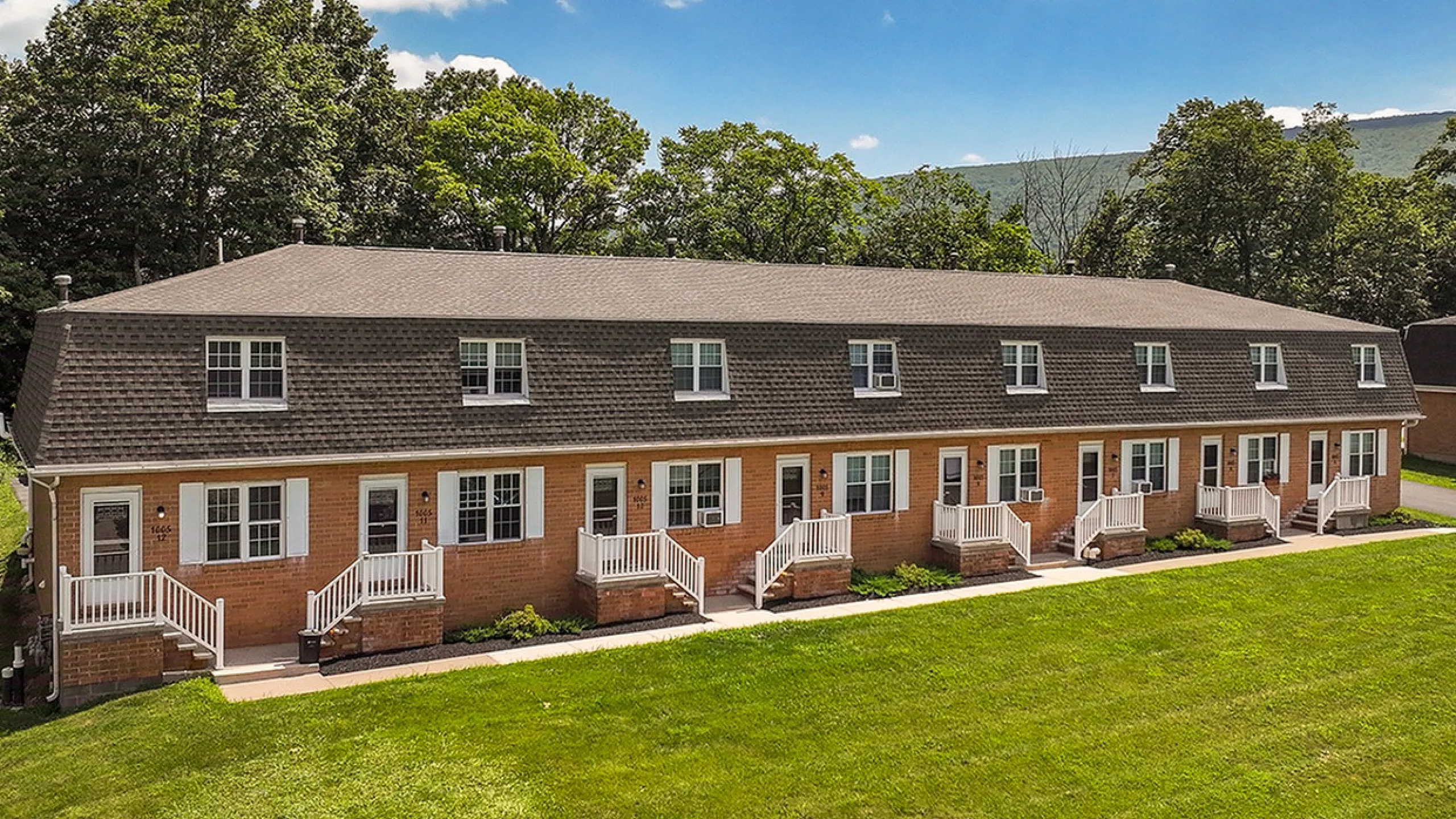 Charming row of red-brick townhouses with picturesque porches, surrounded by lush green lawns and scenic wooded backdrop.