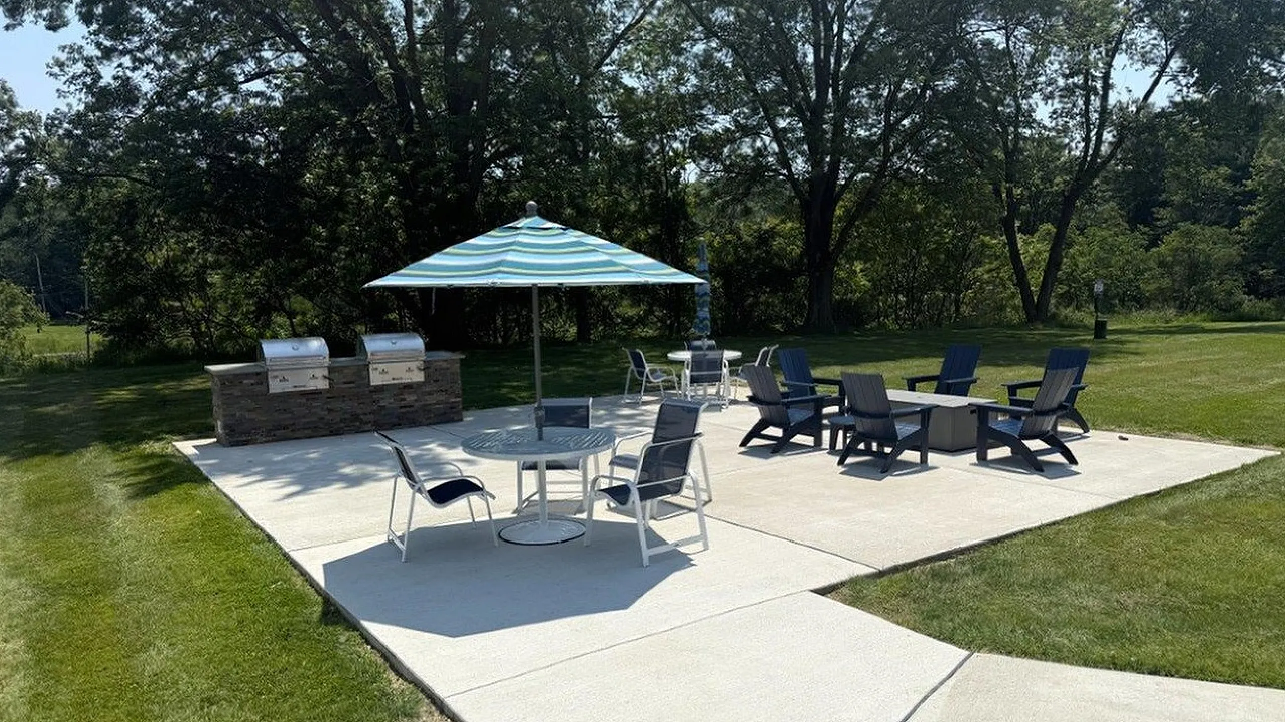 Modern outdoor patio with stylish seating, BBQ grills, and large umbrella, perfect for entertaining in a lush green setting.