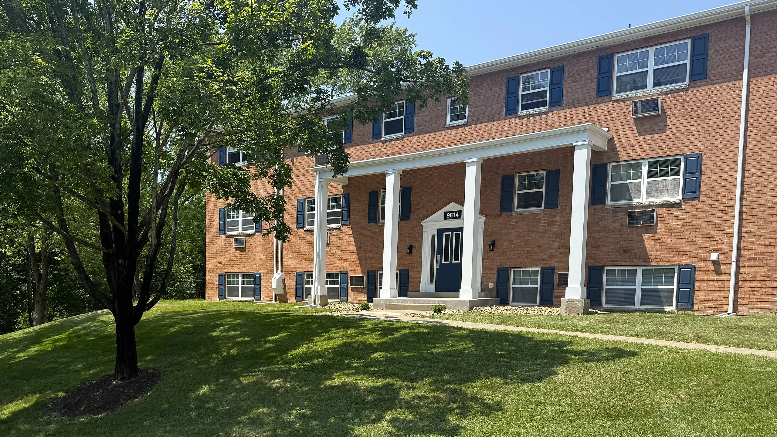 Charming brick apartment building with elegant columns, surrounded by lush green lawns and mature trees on a sunny day.