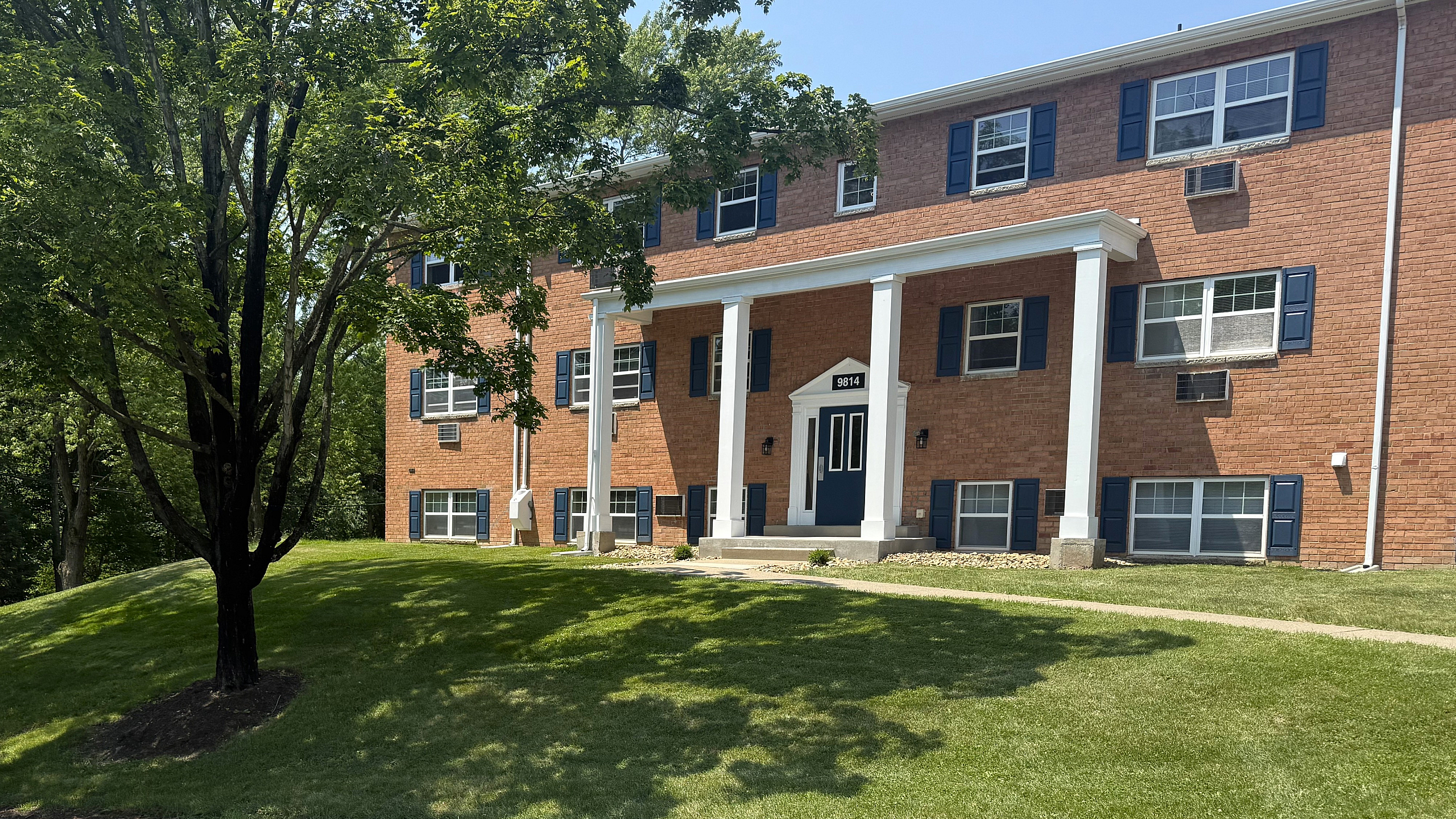 Charming brick apartment building with white columns, nestled in a lush green setting.