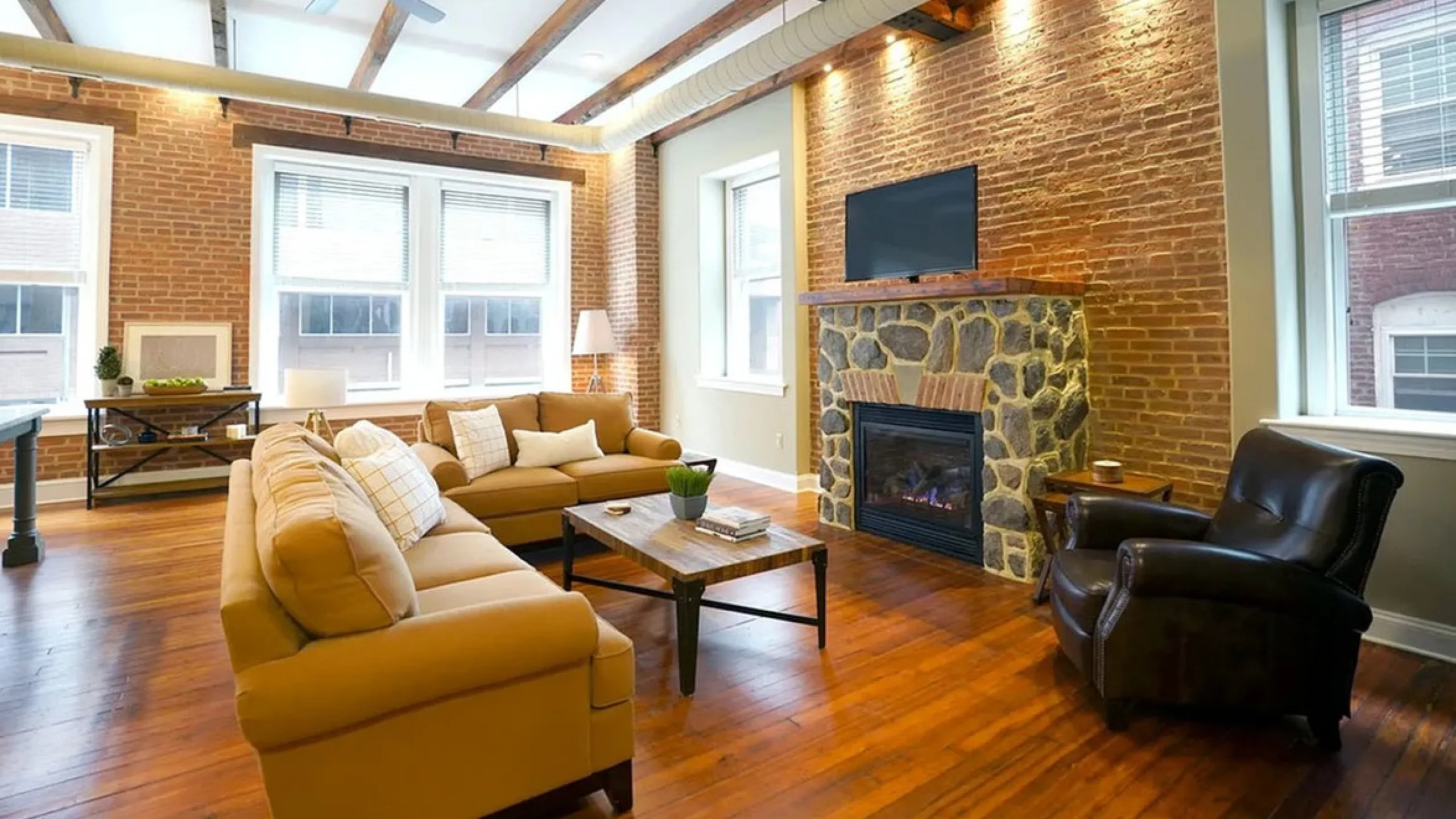 Chic urban living room with rustic exposed brick walls, elegant wooden beams, and cozy fireplace, featuring plush sofas and ample natural light.