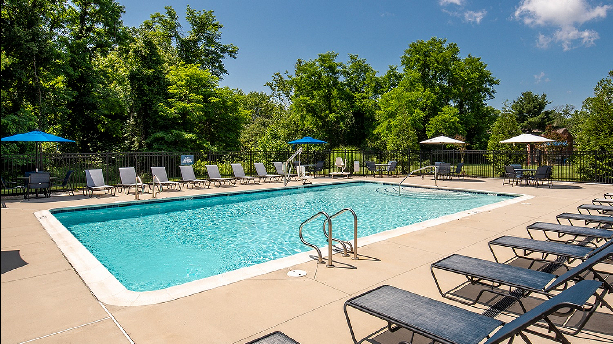 Inviting outdoor pool area surrounded by lush greenery, featuring numerous lounge chairs and shaded tables for relaxation.