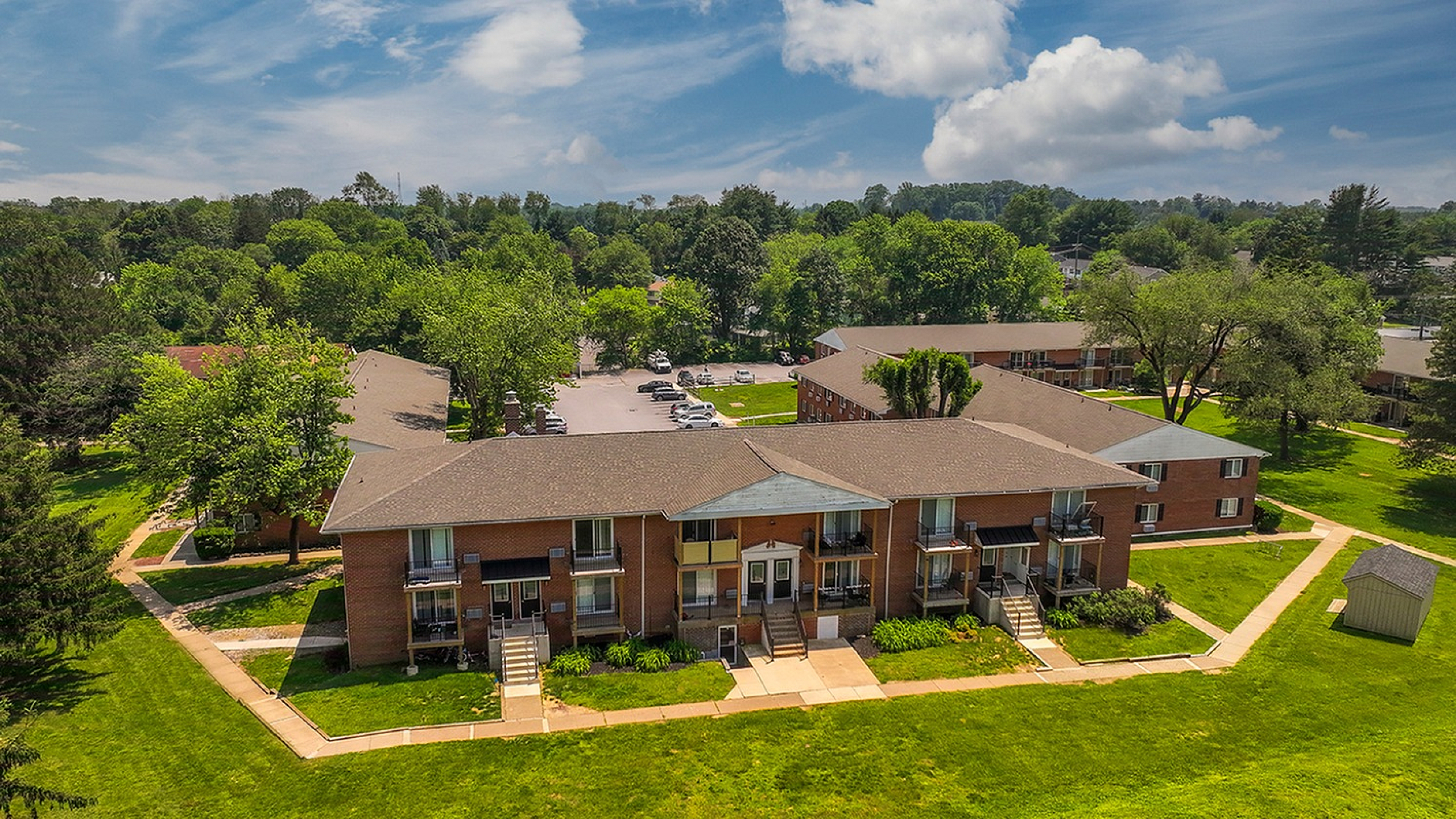Charming apartment complex nestled in a lush, green landscape under a vibrant blue sky, featuring well-maintained brick buildings and spacious grounds.