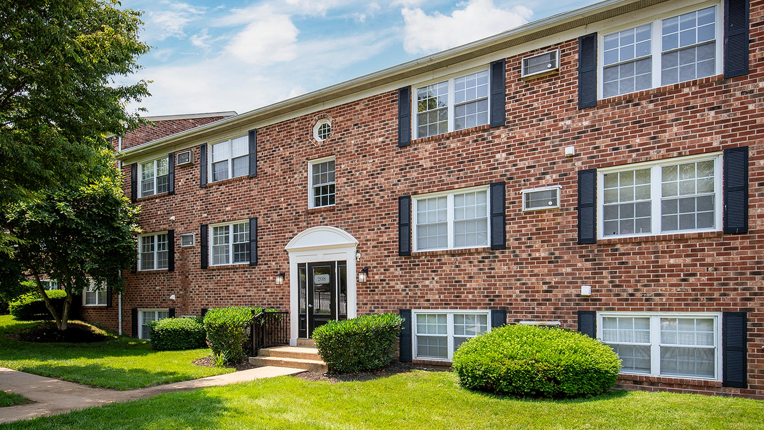 Charming brick apartment building with classic architecture and lush green surroundings, bathed in sunlight for a warm and welcoming feel.