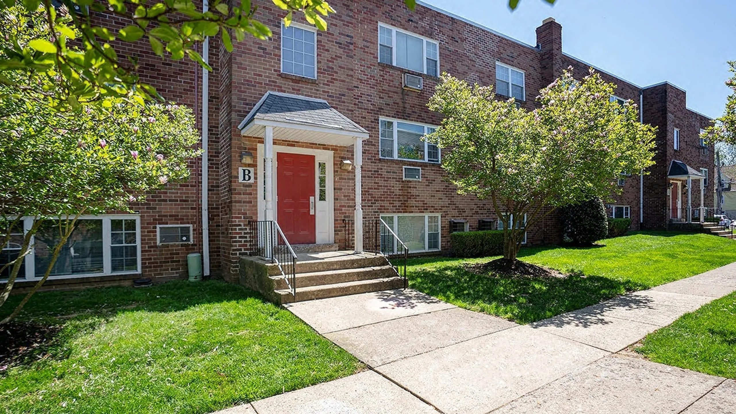 Charming brick apartment building with a welcoming entrance, lush greenery, and sunny surroundings.