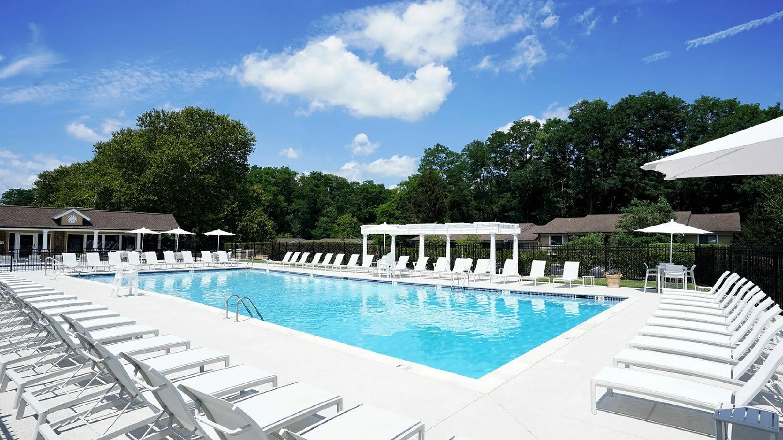 Elegant outdoor swimming pool area with abundant lounge chairs, surrounded by lush greenery and a bright blue sky, offering a perfect relaxation spot.