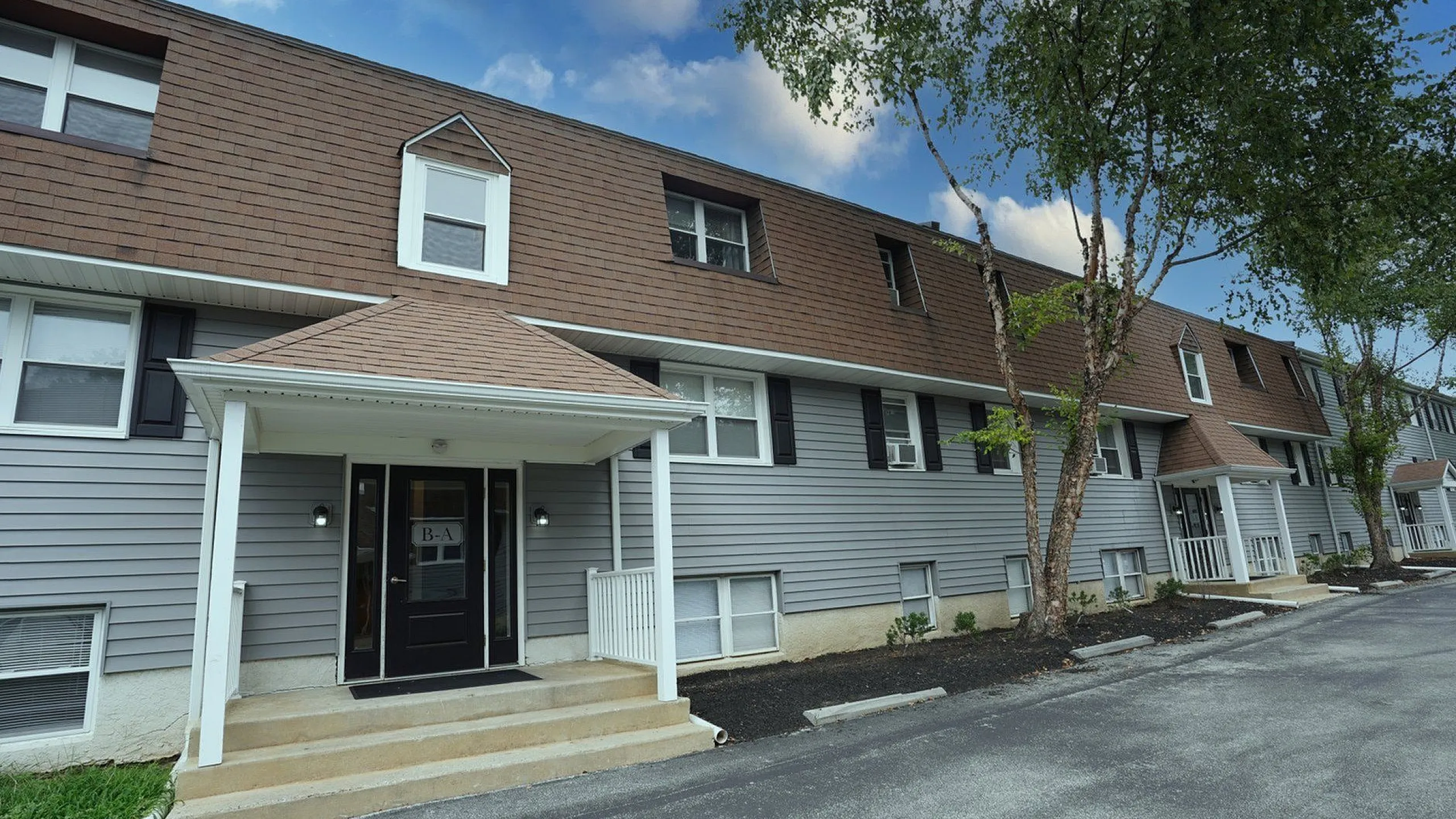 Charming townhouse-style apartment with a modern facade, featuring elegant brown and gray exterior, welcoming porch, and picturesque trees in a serene setting.