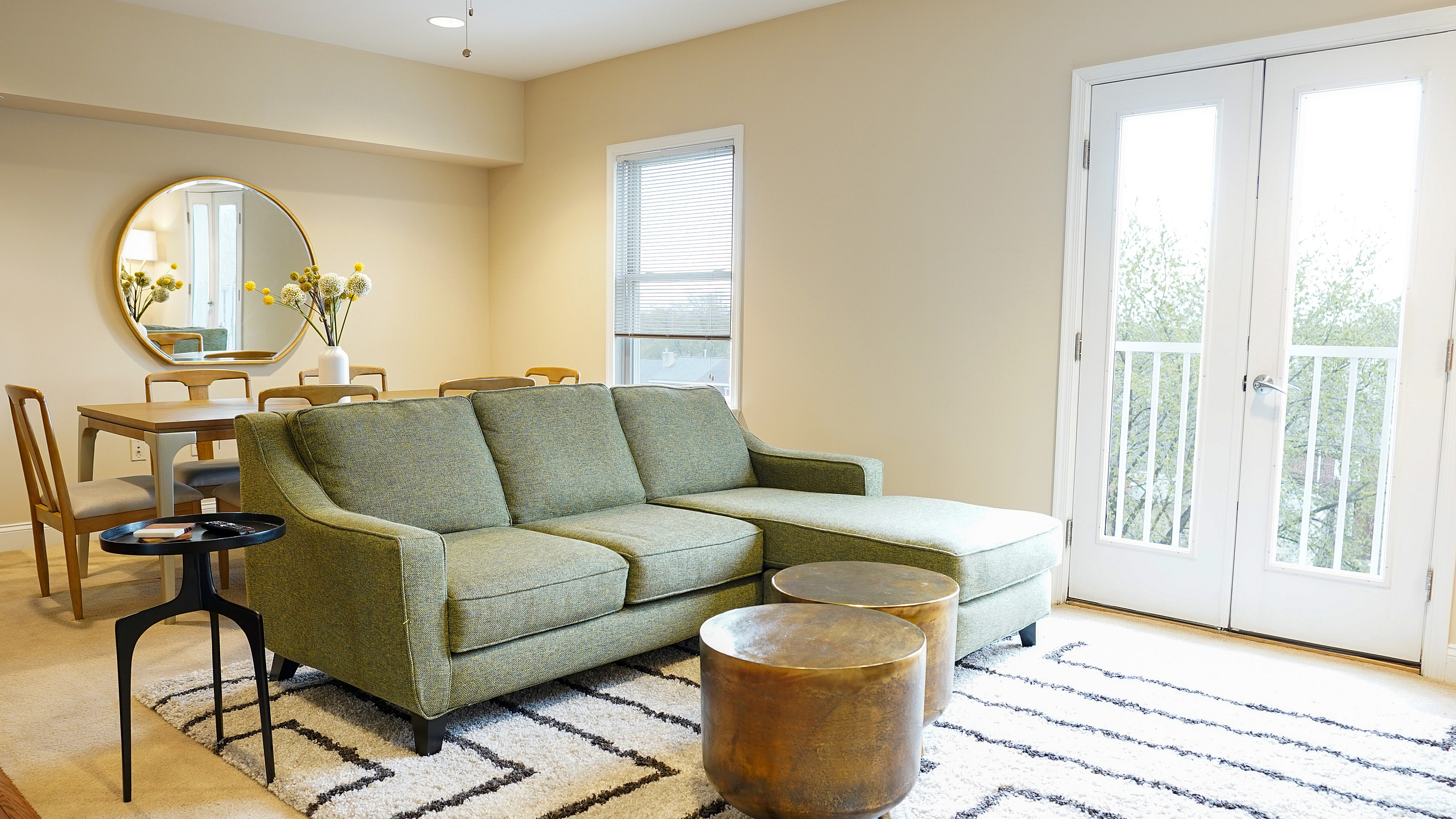 Bright and cozy living room featuring a stylish green sectional sofa, elegant round coffee tables, and a chic dining area by the window. Natural light floods through large glass doors, highlighting the modern decor.