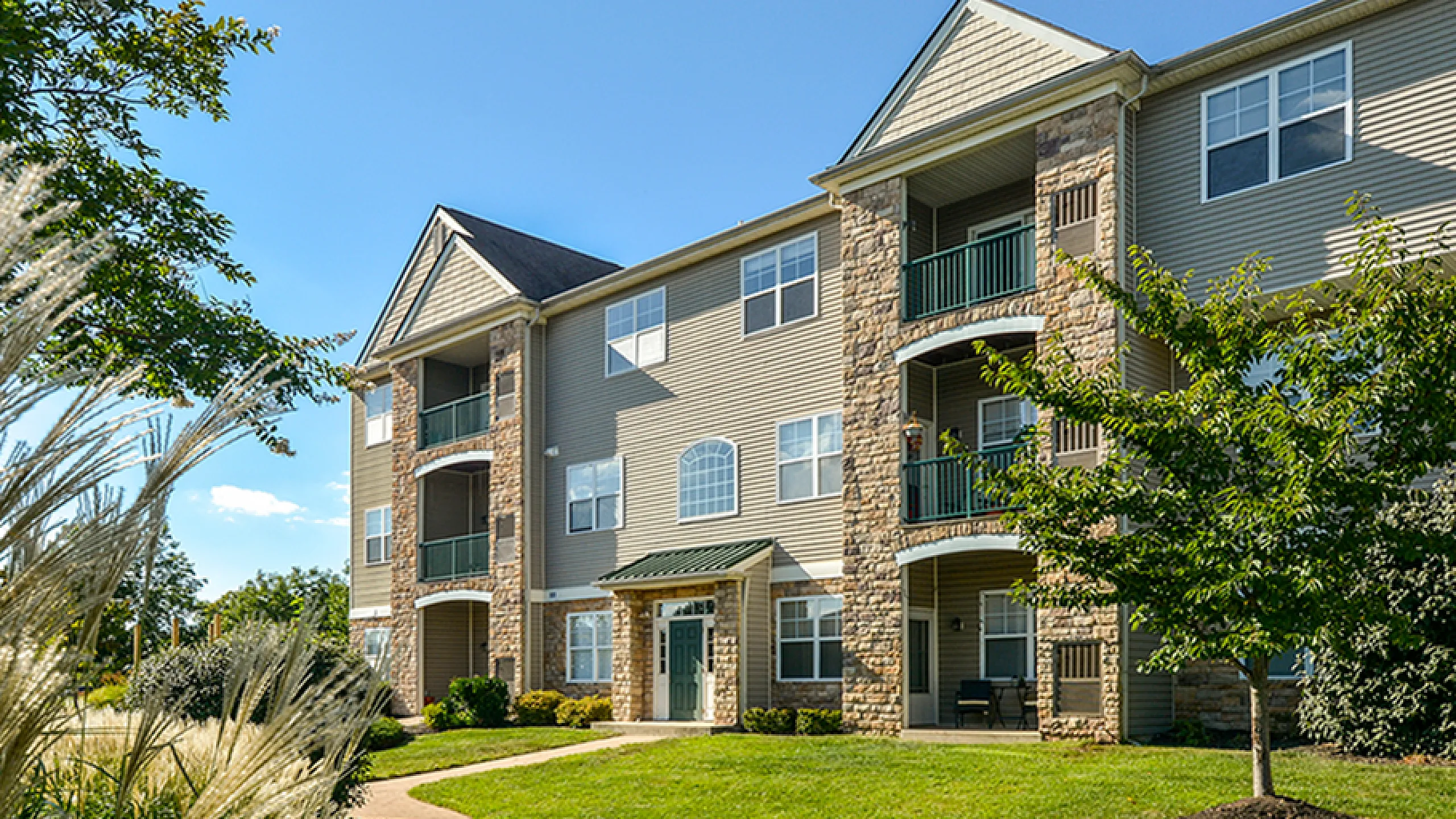 Charming apartment building with stone accents and lush greenery on a sunny day, showcasing inviting balconies and a well-kept lawn.