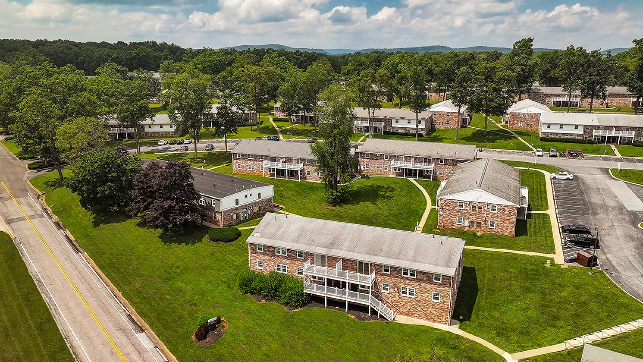 Charming aerial view of a beautifully landscaped apartment community with brick buildings, lush green lawns, and scenic surroundings.
