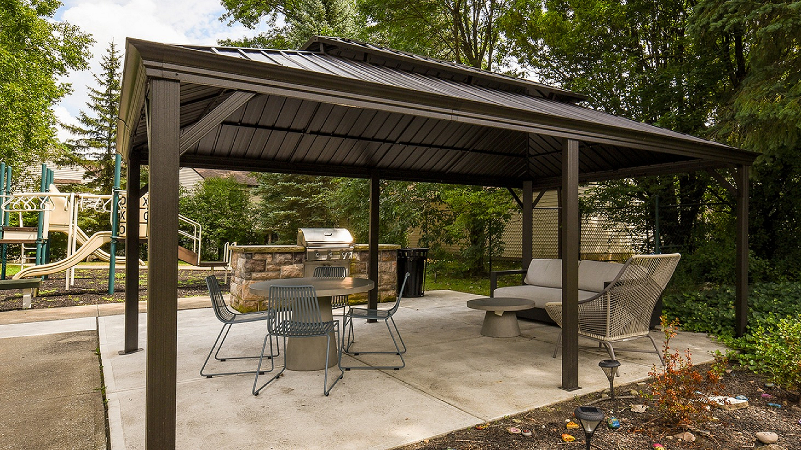 Inviting outdoor space featuring a modern gazebo with comfortable seating and a built-in grill, perfect for entertaining or relaxing amidst lush greenery.