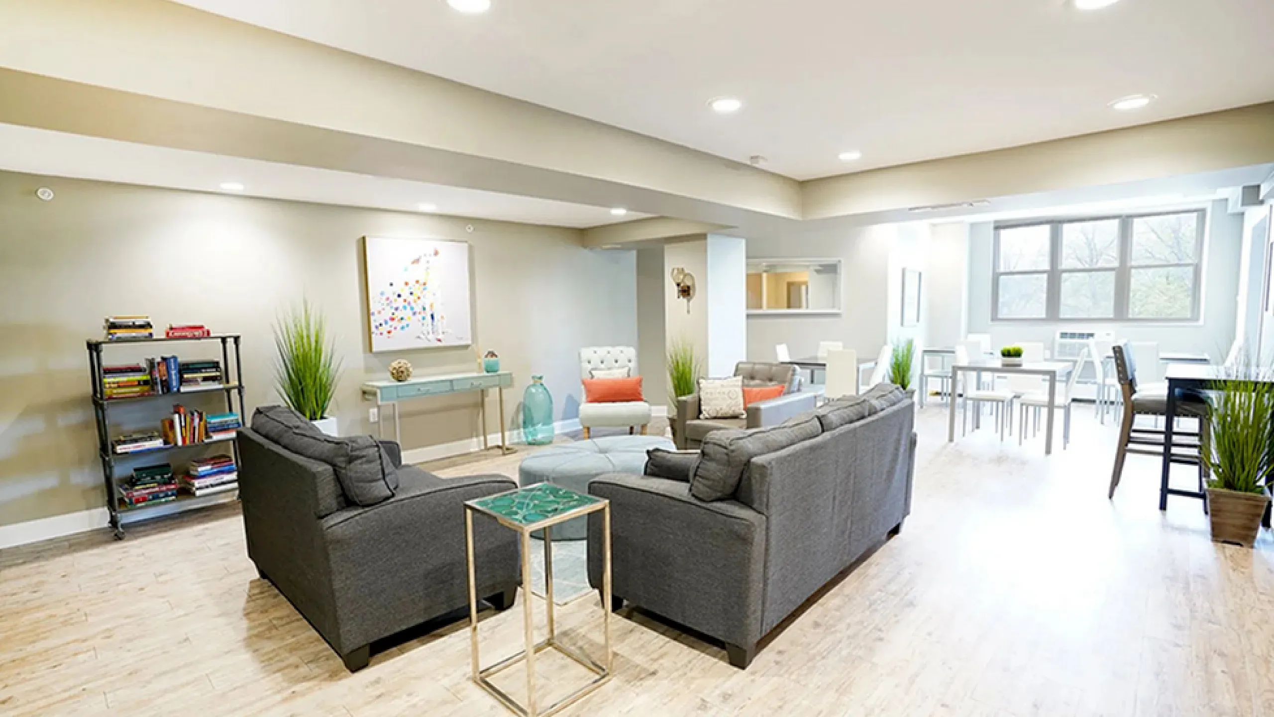 Bright and spacious modern living area with cozy gray sofas, vibrant decor, and an inviting dining space featuring large windows and lush greenery.