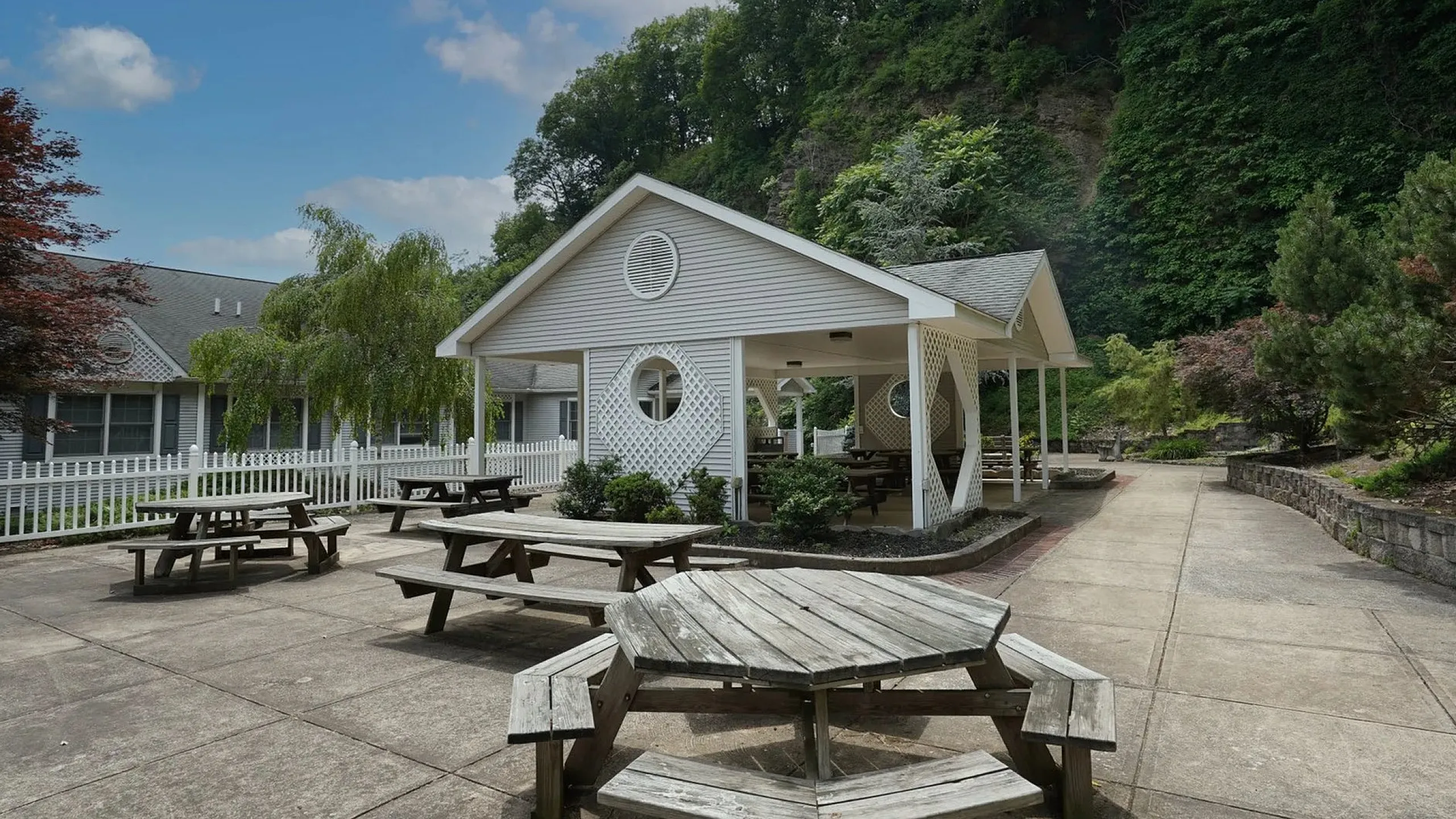 Charming outdoor patio area with wooden picnic tables, nestled beside lush greenery and a quaint pavilion, perfect for gatherings.