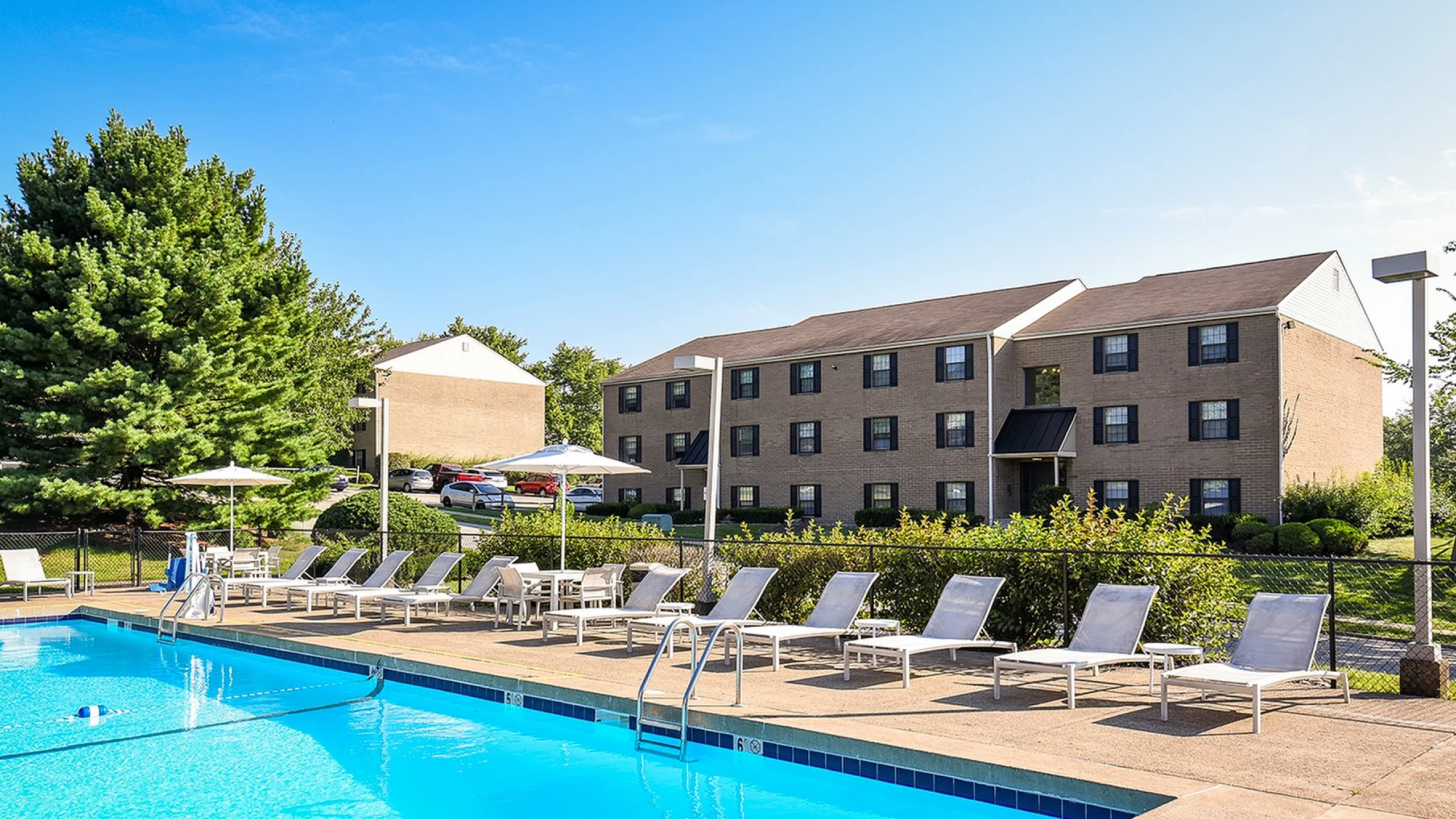Inviting apartment complex with a crystal-clear swimming pool and relaxing lounge chairs, surrounded by lush greenery under a clear blue sky.