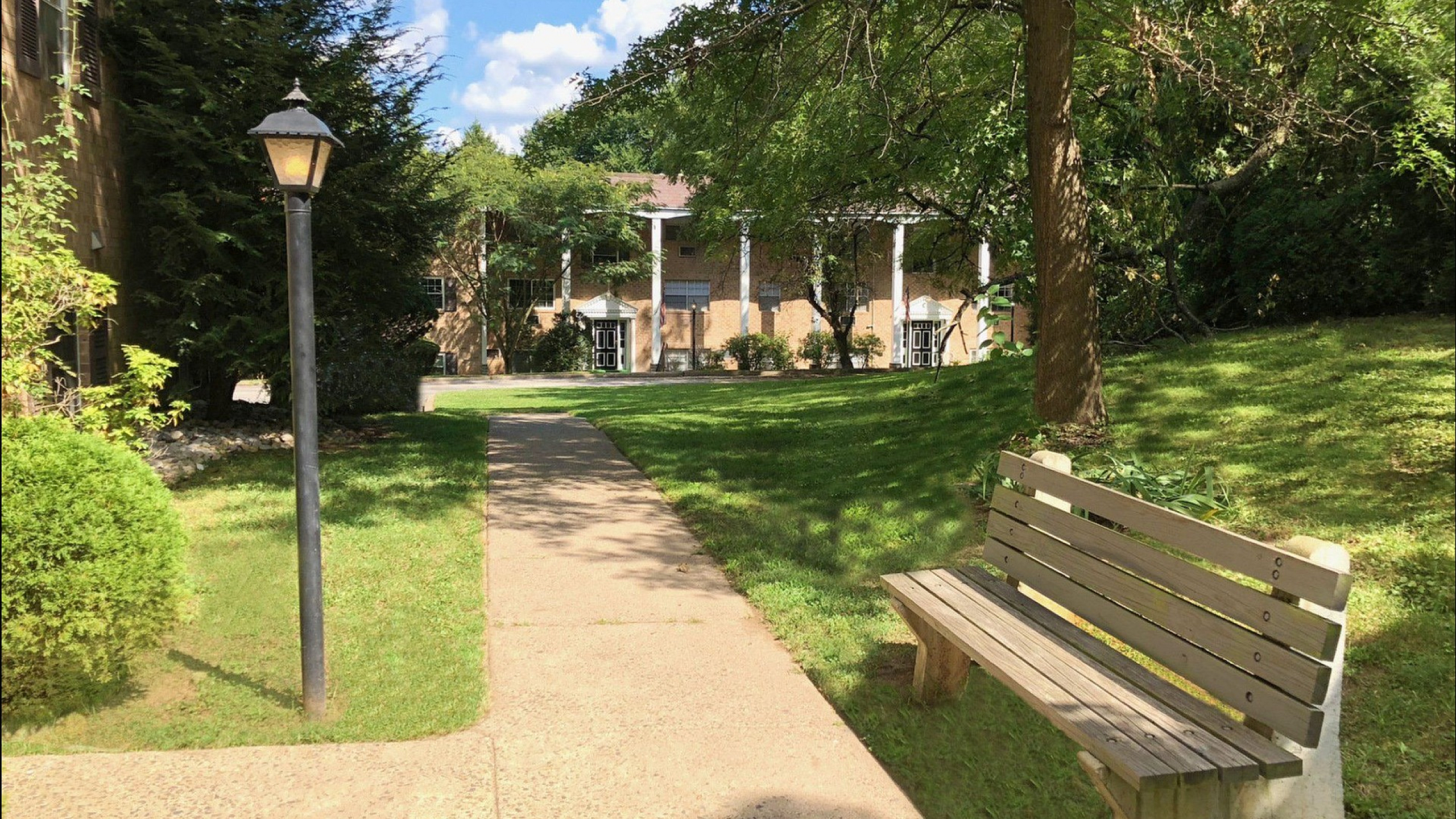 Charming garden pathway with a wooden bench leading to elegant apartment entrance, surrounded by lush greenery.