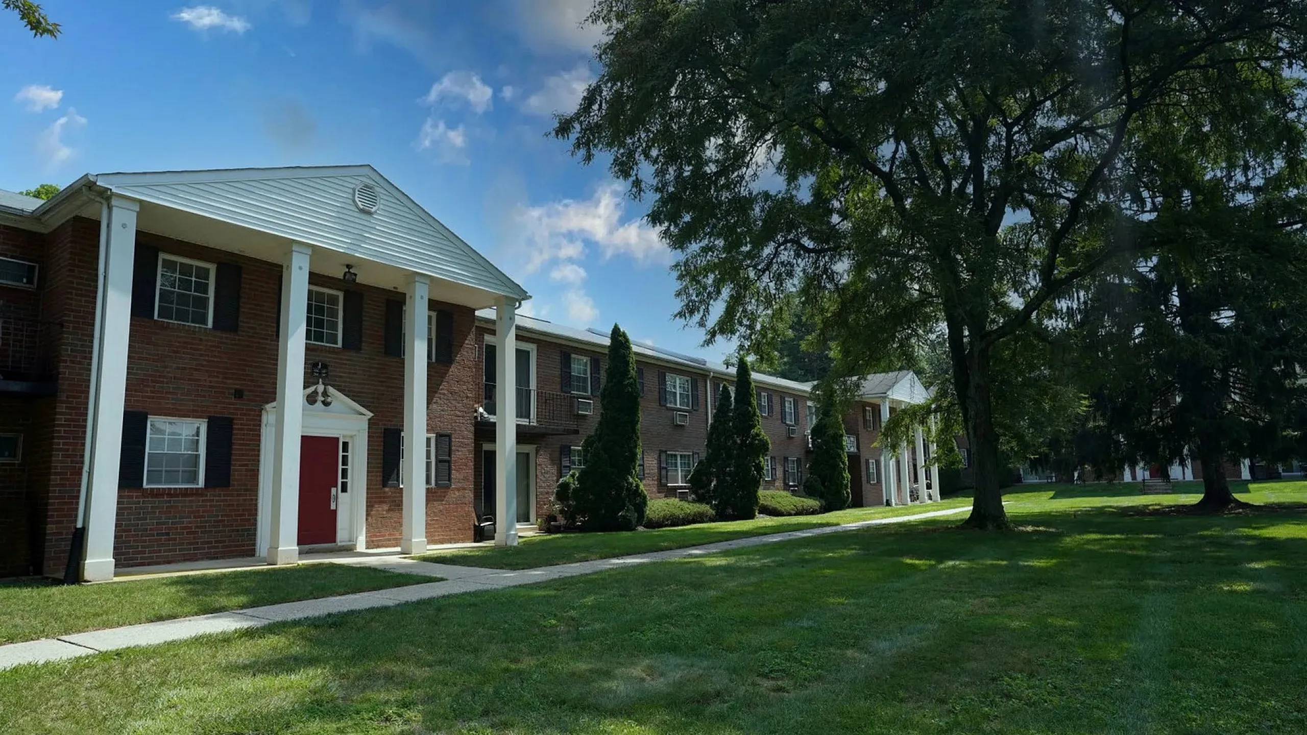 Charming red brick apartment complex with classic white column entrances and lush green lawns under a bright blue sky.