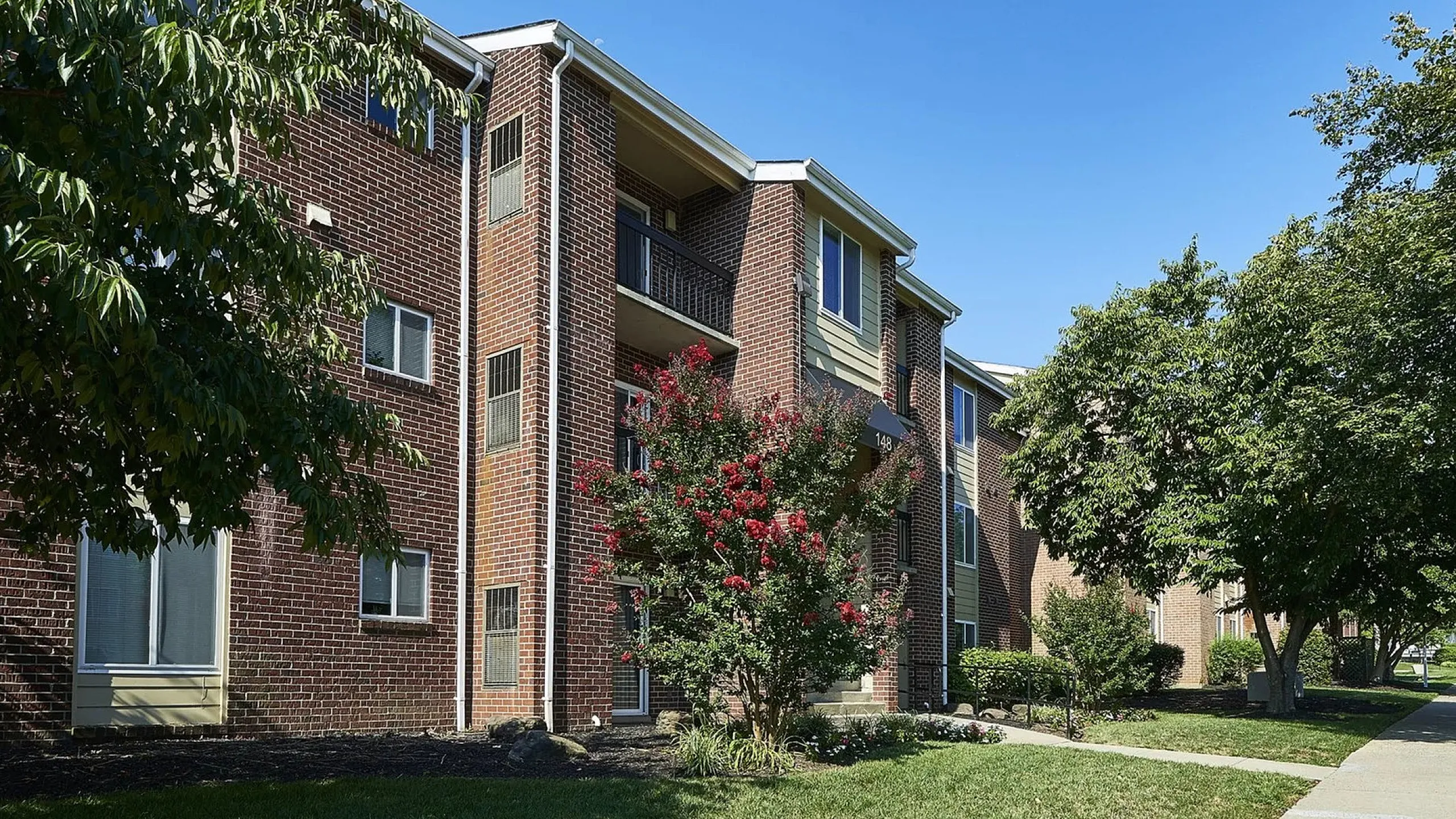 Charming brick apartment complex surrounded by lush greenery and vibrant red blossoms, offering a serene and inviting residential environment.