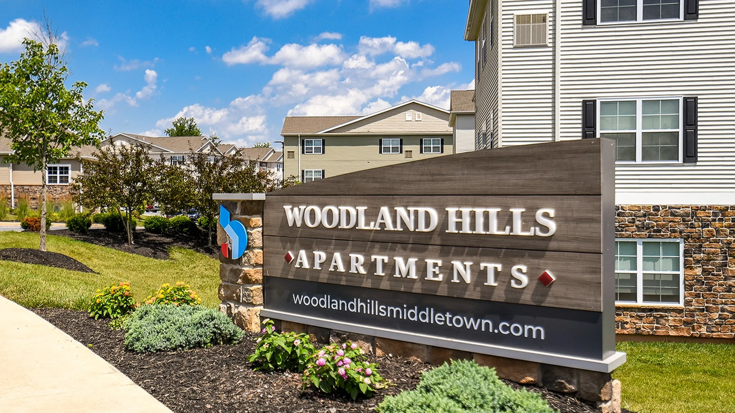 Charming Woodland Hills Apartments sign amidst vibrant landscaping and modern residential buildings under a bright blue sky.