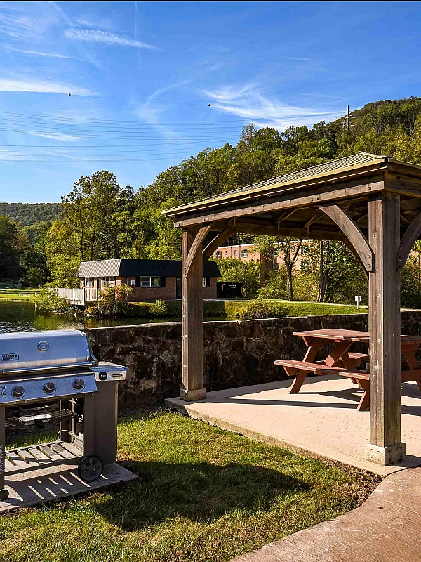 Charming outdoor pavilion with a wooden pergola and picnic table, overlooking scenic water and lush greenery, ideal for serene outdoor gatherings.