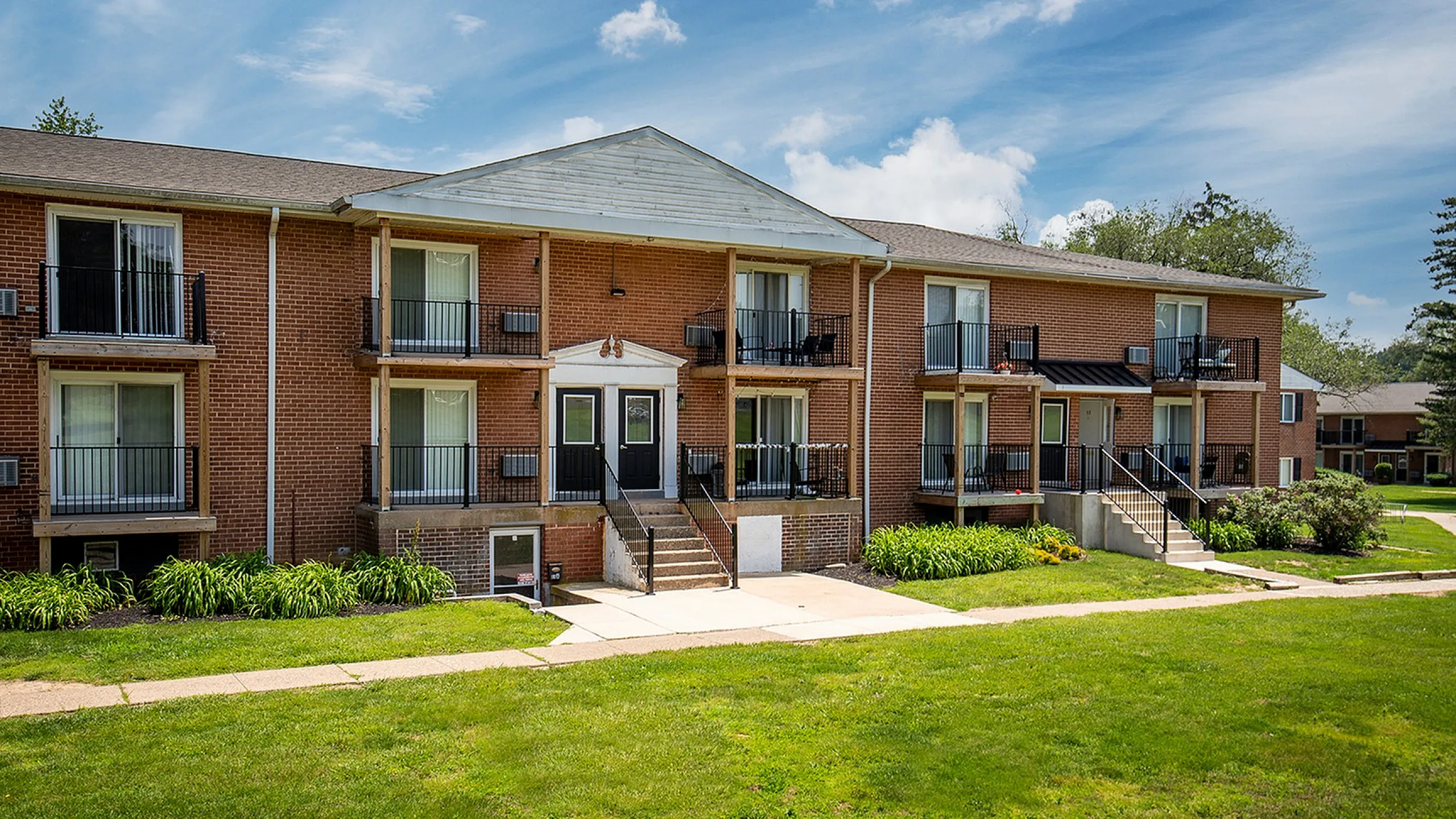 Charming apartment building with red brick facade, small balconies, and lush green lawns under a clear blue sky.