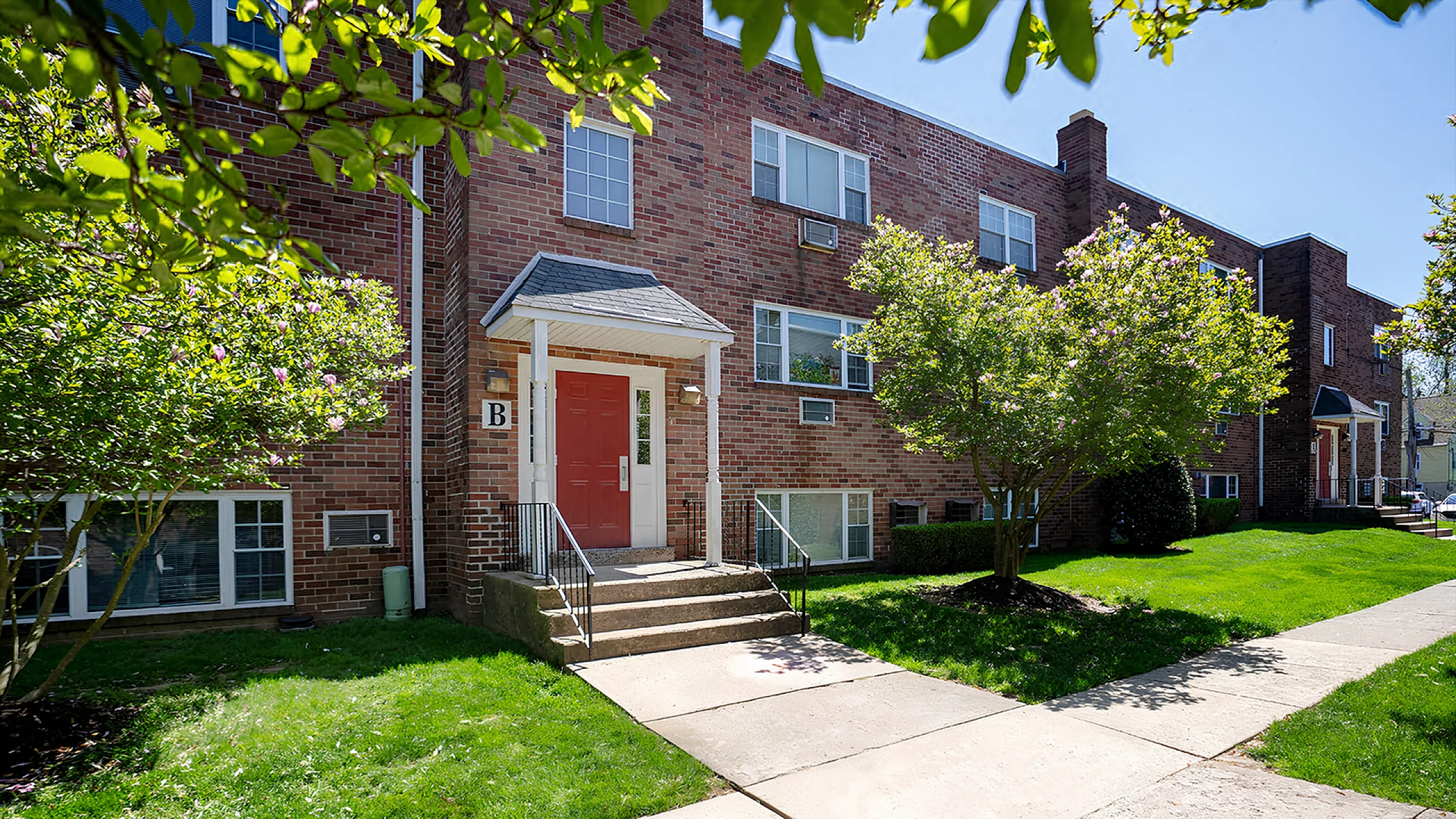 Inviting brick apartment building with lush green lawns and charming red door entrance, perfect for modern living.