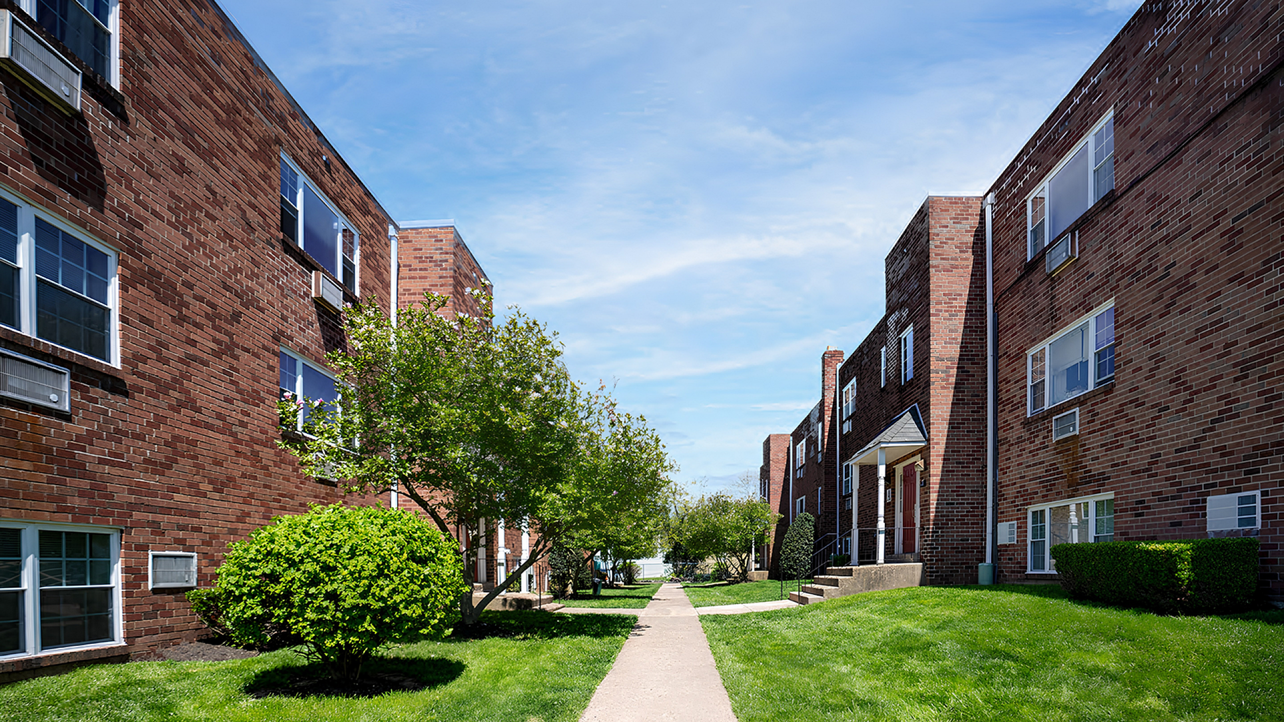 Charming apartment complex with classic brick architecture, lush green landscaping, and a serene pathway.