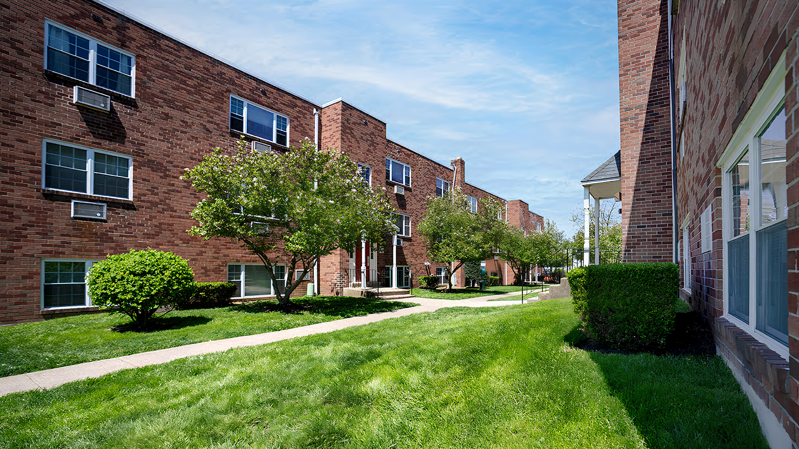 Charming brick apartment complex with lush green lawns, mature trees, and large windows under a clear blue sky.