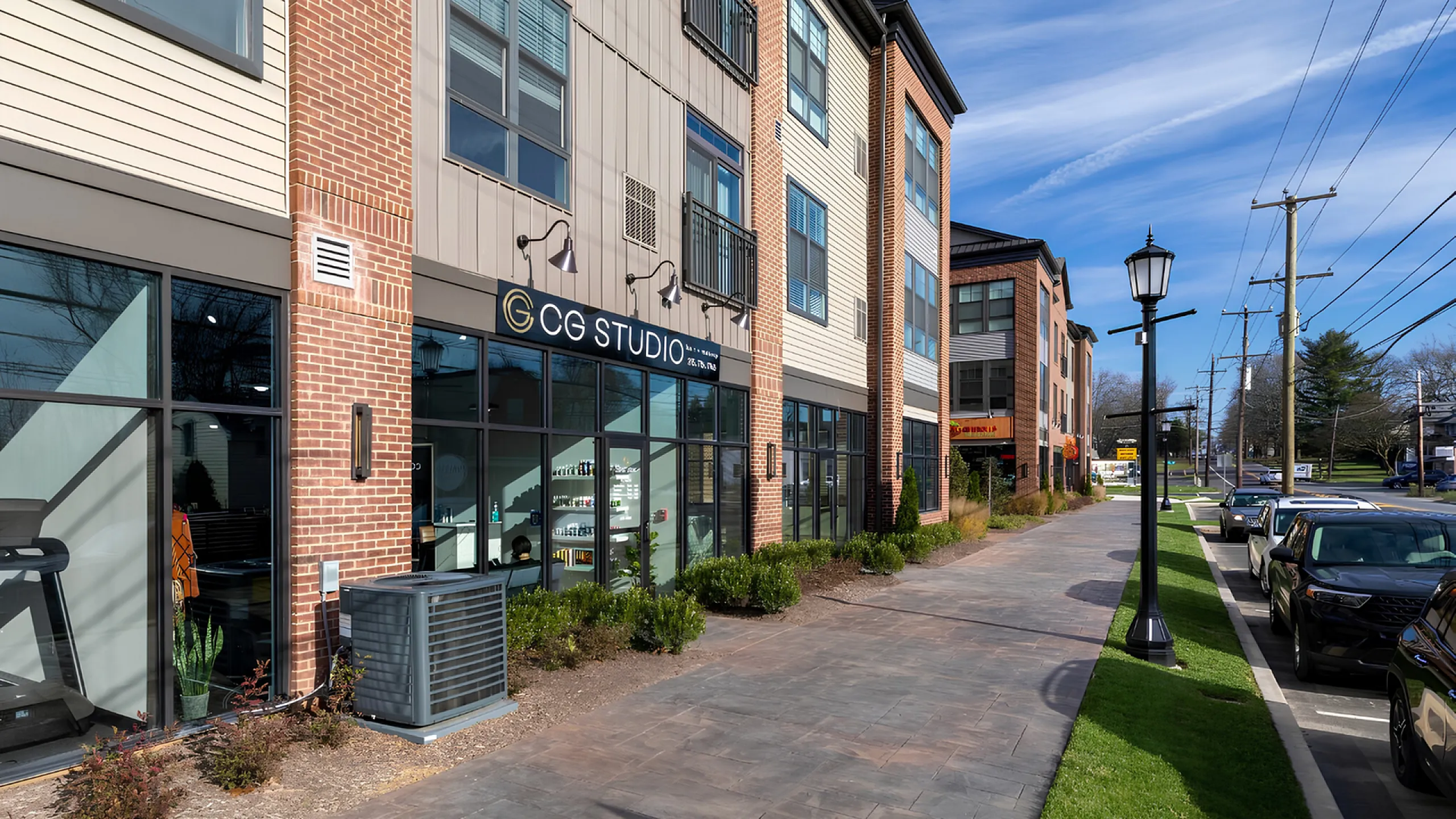 Charming urban streetscape featuring a modern brick apartment building with ground-floor retail space, surrounded by lush greenery and ample parking.