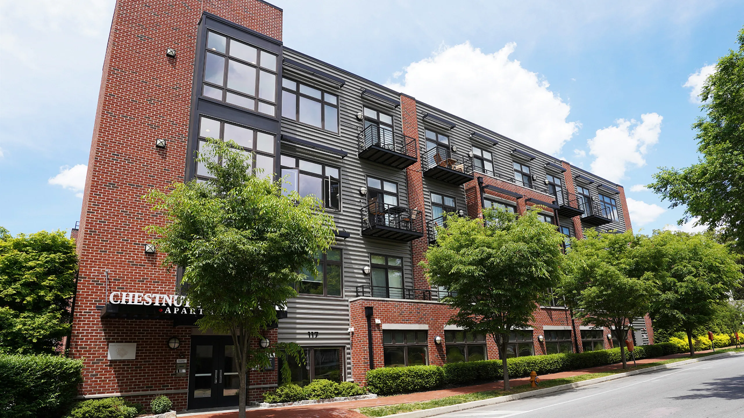 Charming brick apartment building with contemporary windows, lush greenery, and inviting balconies, set against a clear blue sky.