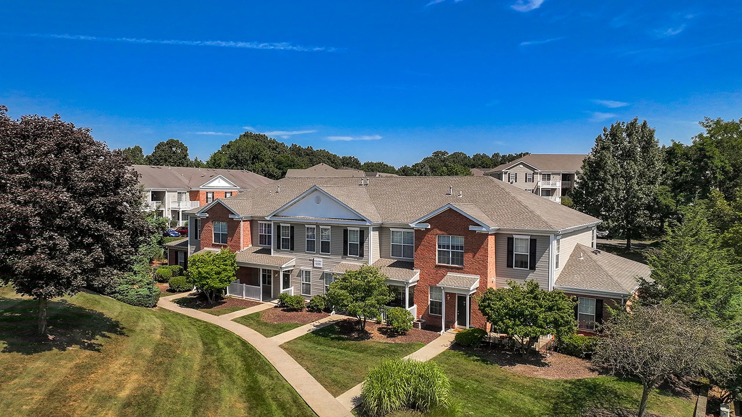 Charming brick apartment complex surrounded by lush greenery and clear blue skies, featuring well-maintained walkways and inviting entryways.