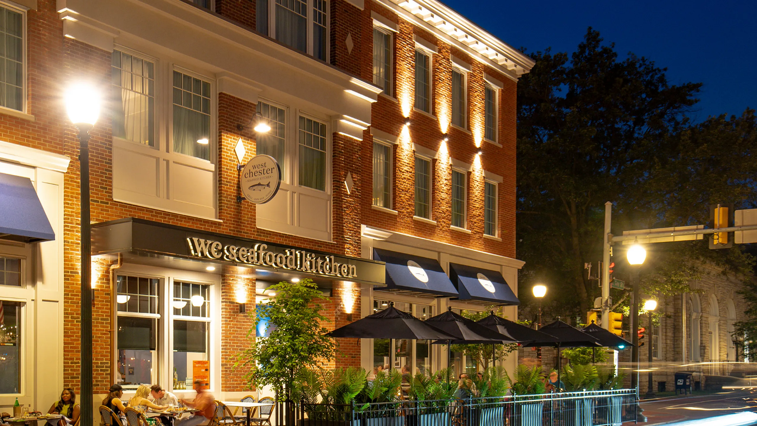 Charming street view of a brick building with inviting outdoor seating at a seafood kitchen, glowing warmly under ambient evening lights.