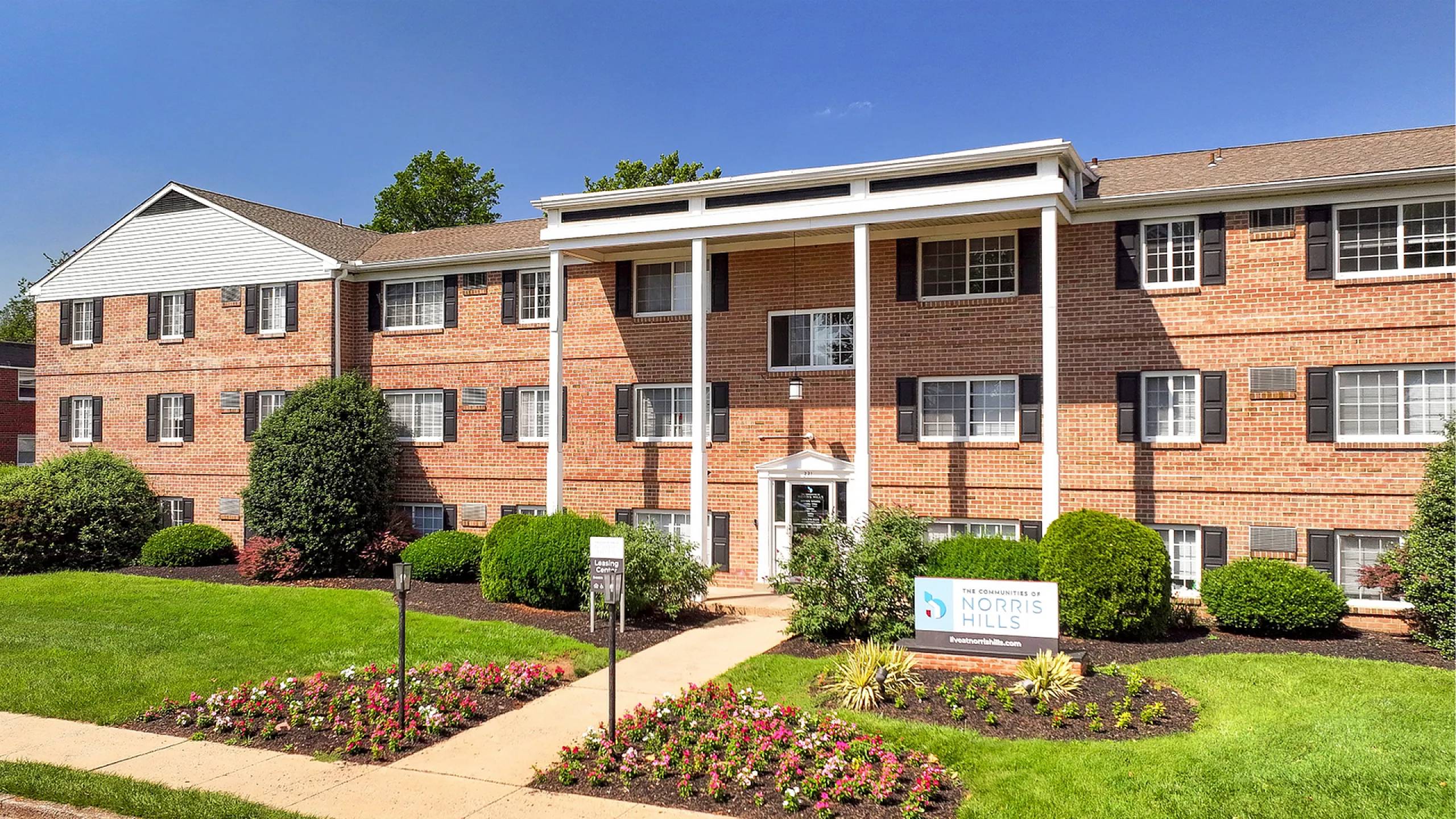 Charming red-brick apartment building with colonial-style pillars, surrounded by lush landscaping and vibrant flower beds, perfect for stylish and comfortable living at Norris Hills.