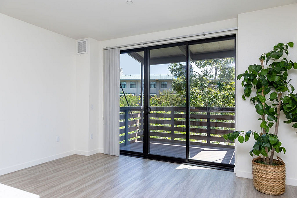 Bright and airy room with elegant wood flooring, floor-to-ceiling sliding glass doors leading to a scenic balcony, and lush greenery for a serene ambiance.