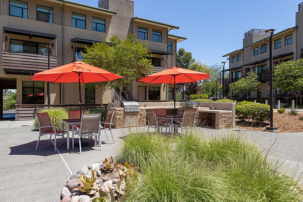 Elegant courtyard with vibrant orange umbrellas, outdoor seating, and grilling station surrounded by lush greenery.
