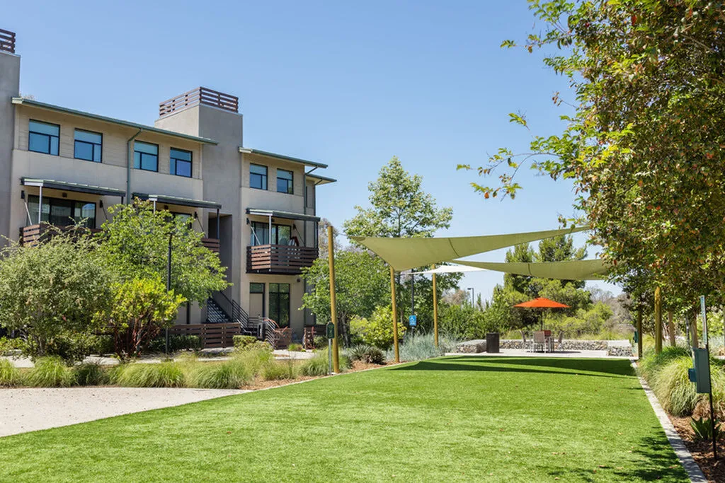 Modern apartment building surrounded by lush greenery and a spacious pet park area featuring sunshades and seating.