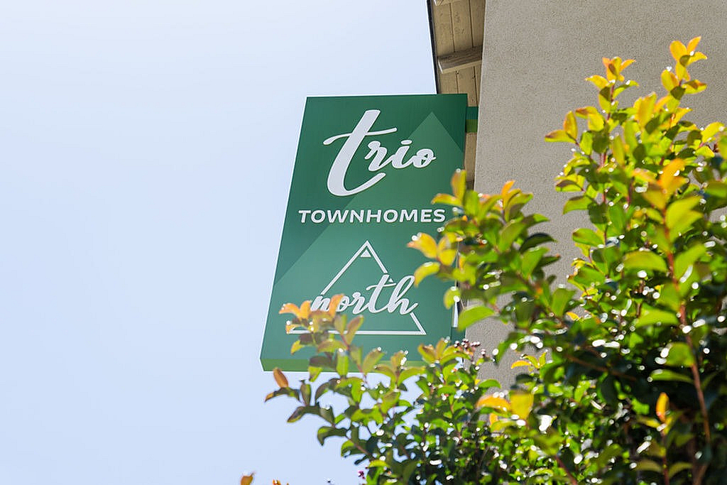 Inviting green sign for Trio Townhomes North against clear sky, surrounded by vibrant green foliage.