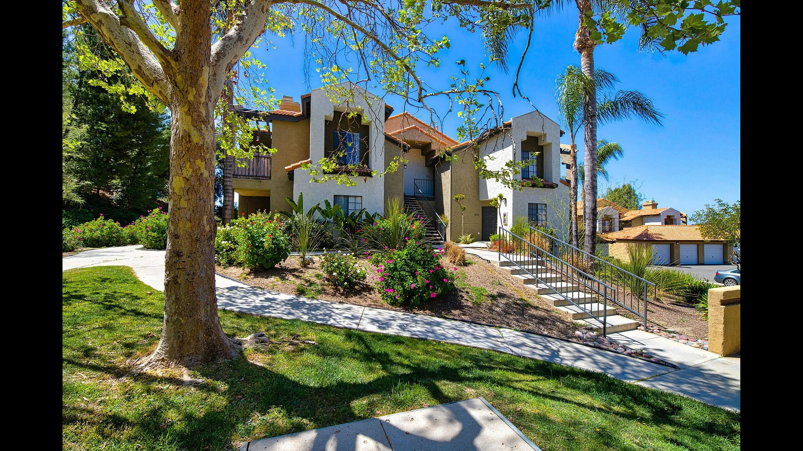 Charming apartment complex with lush green landscaping, inviting pathways, and vibrant foliage, set under a bright, clear blue sky.