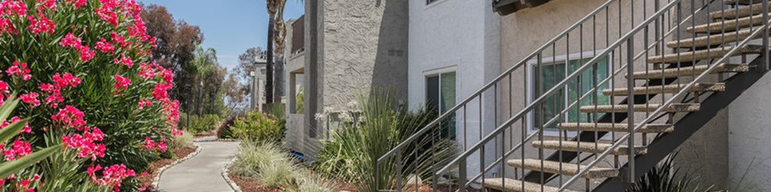 Charming apartment building surrounded by vibrant pink flowers and lush greenery, with elegant exterior stairs and a welcoming pathway, perfect for nature lovers.