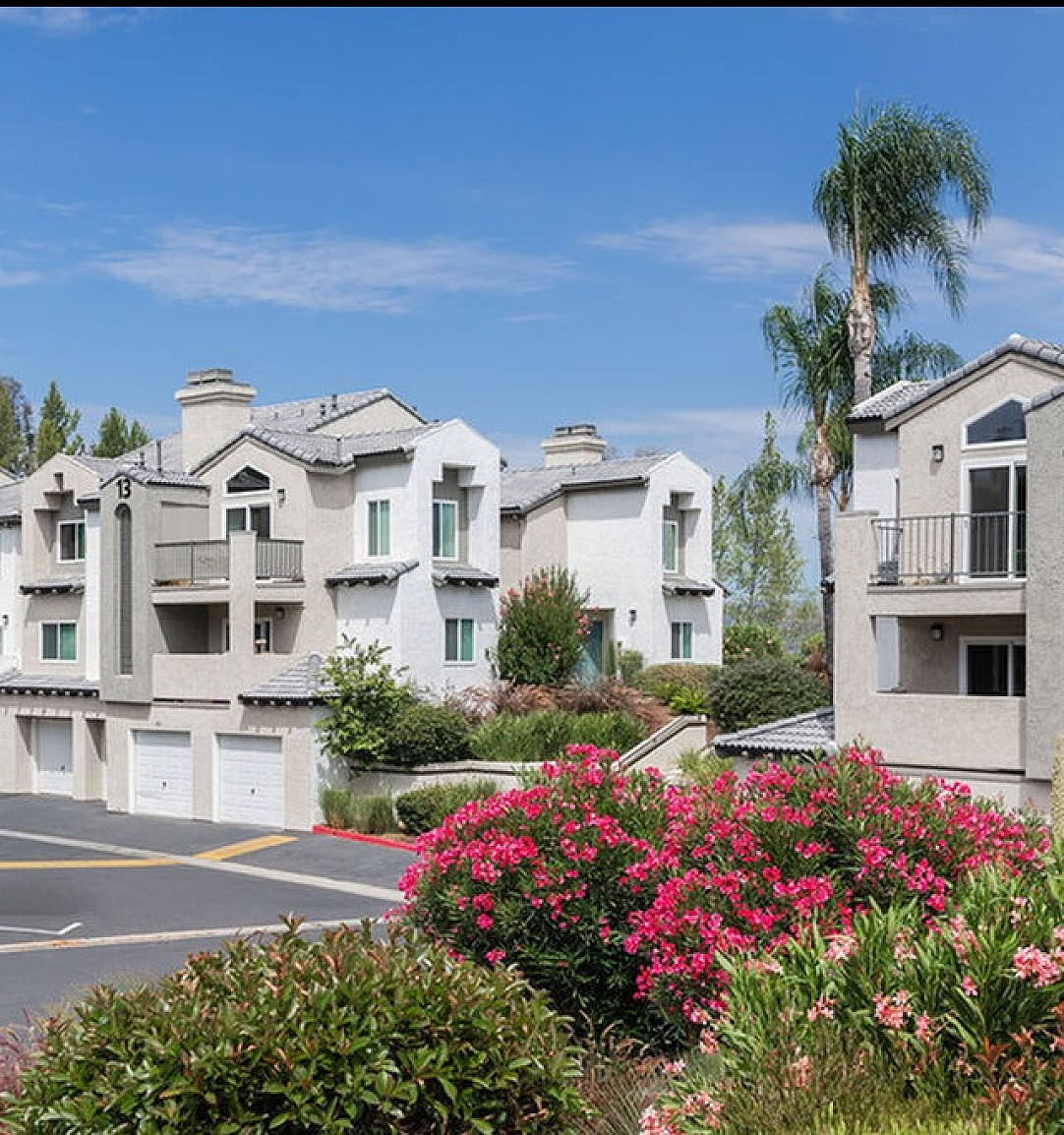 Charming modern apartments with beautiful balconies, surrounded by lush greenery and vibrant flowers under a clear blue sky.