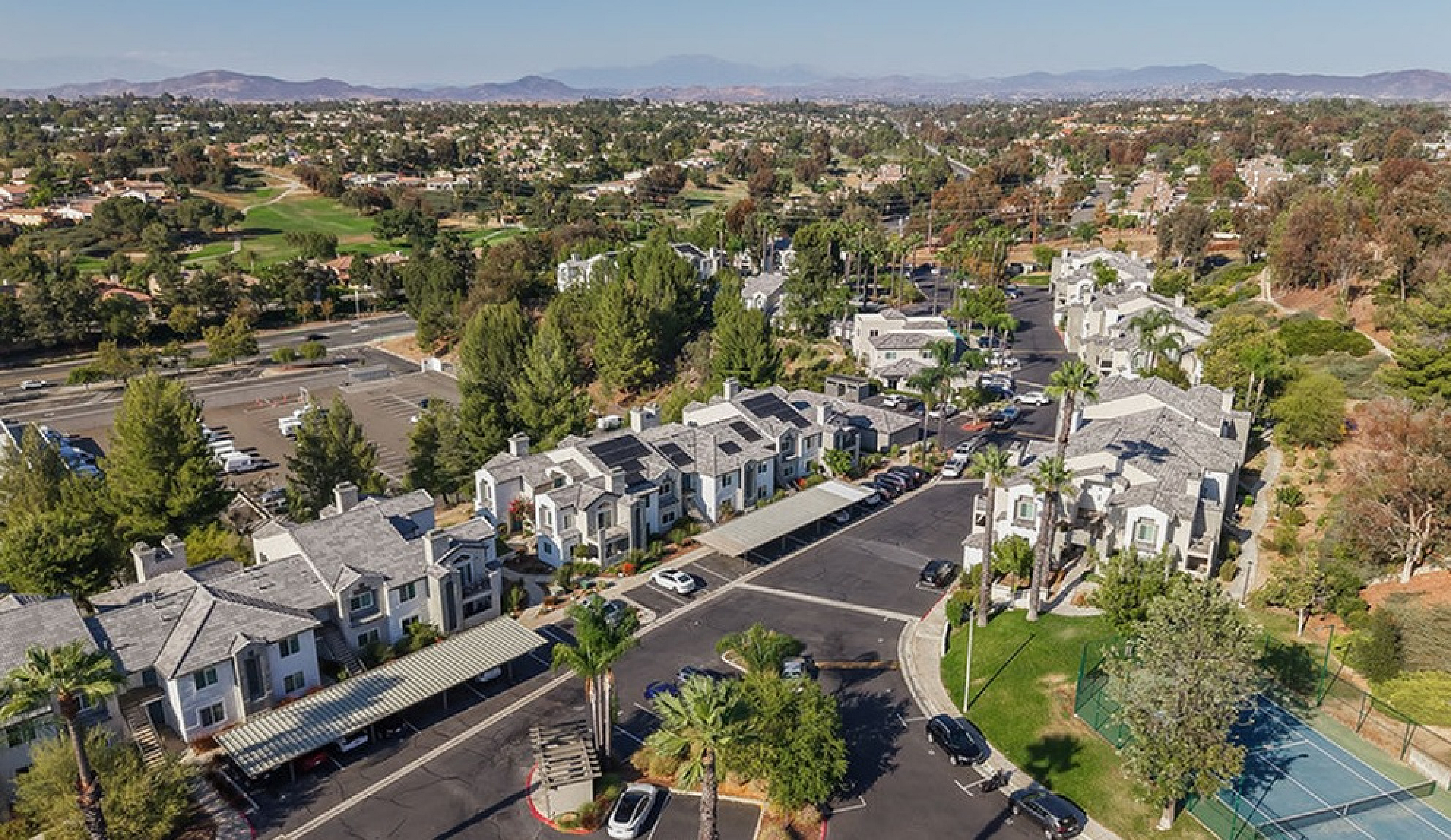 Charming aerial view of a picturesque residential community with beautifully designed townhouses, nestled beside lush greenery and a tennis court, offering serene living surrounded by nature.