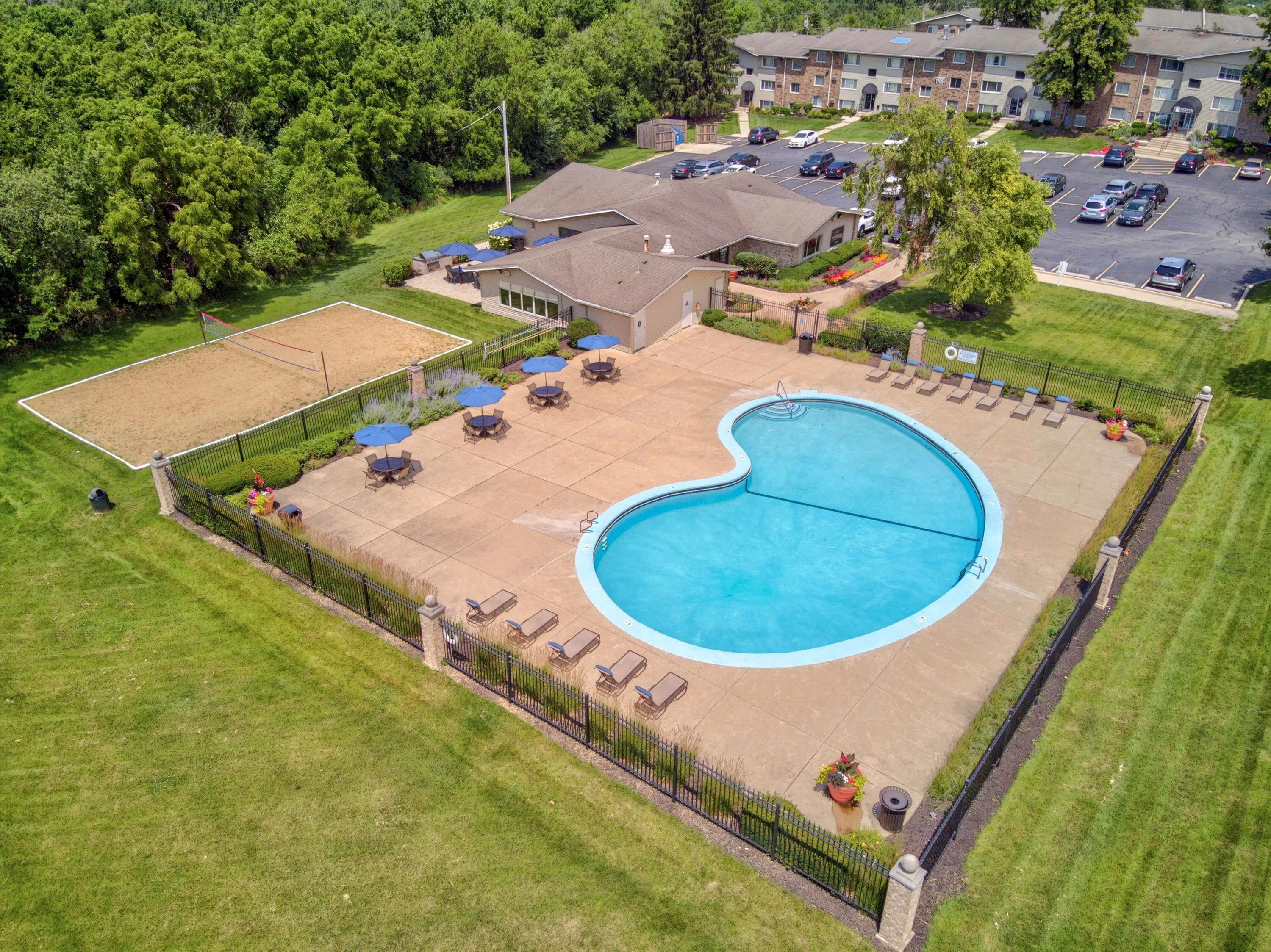 Aerial view of a vibrant residential complex showcasing a heart-shaped swimming pool, expansive sun deck with loungers and umbrellas, adjacent volleyball court, and lush green surroundings.