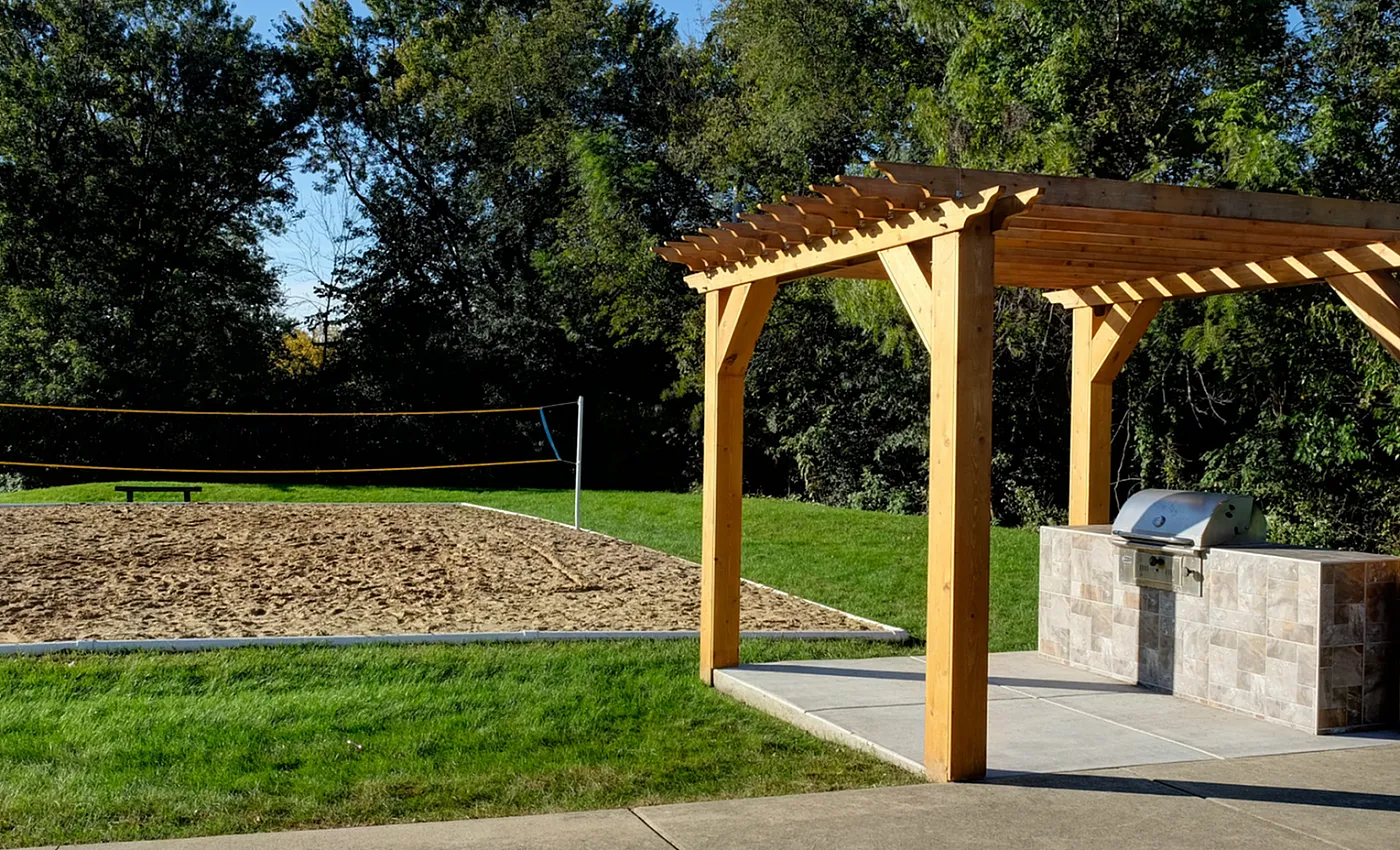 Charming outdoor living area with a wooden pergola and built-in grill beside a sandy volleyball court, surrounded by lush greenery.