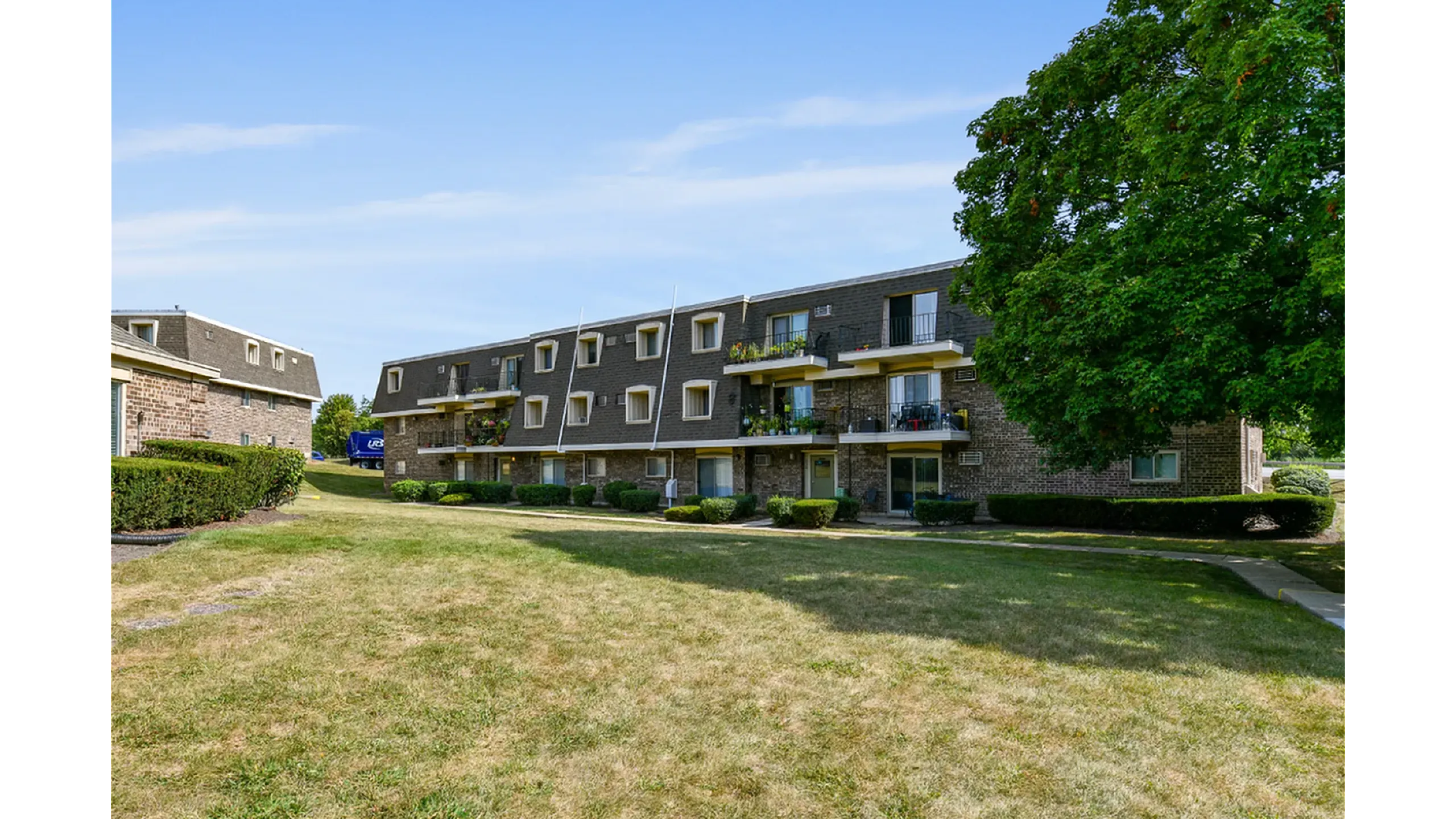 Charming brick apartment building with balconies, surrounded by lush greenery and a large front lawn, set against a bright blue sky.