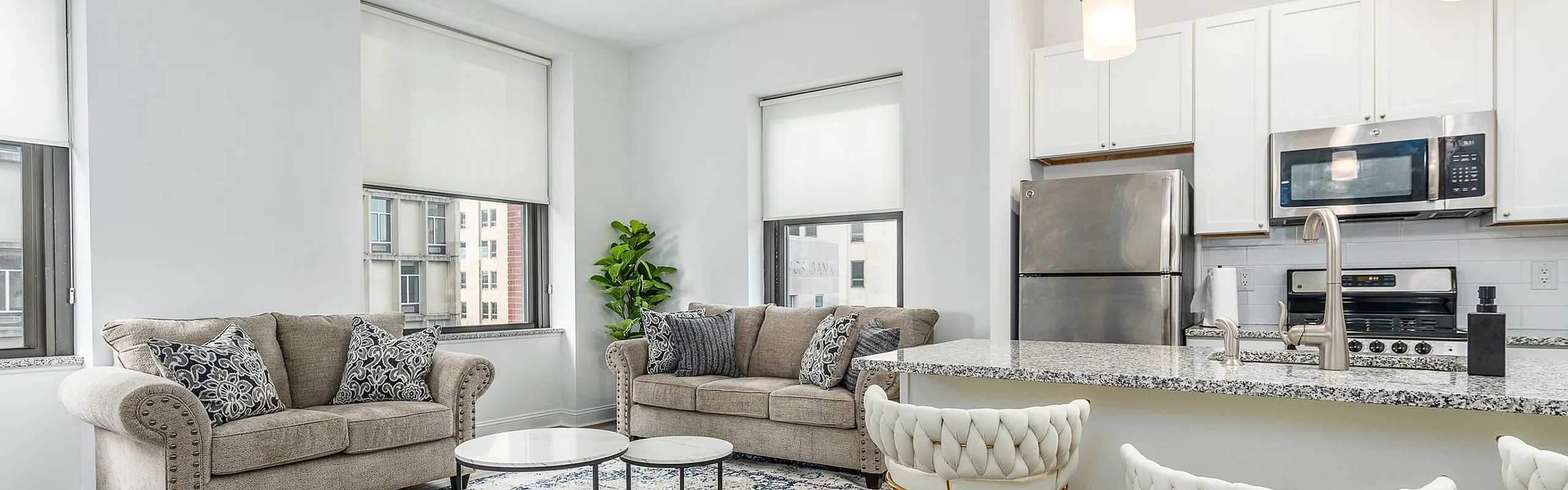Modern open-concept living area with stylish sofas, elegant kitchen, and chic bar seating, bathed in natural light from large windows.